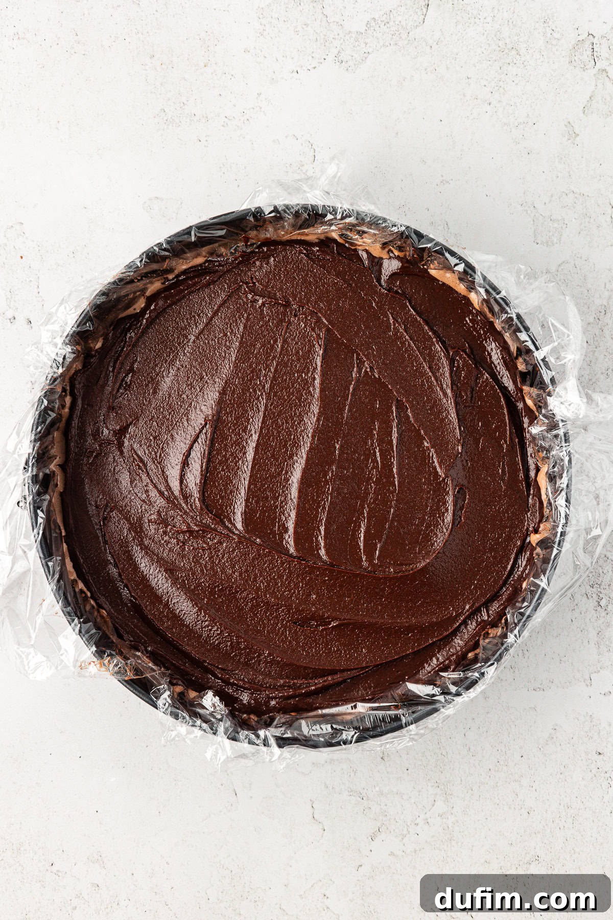 A perfectly layered ice cream cake in a plastic-lined springform pan, with the chocolate fudge ganache evenly spread over the chocolate ice cream.