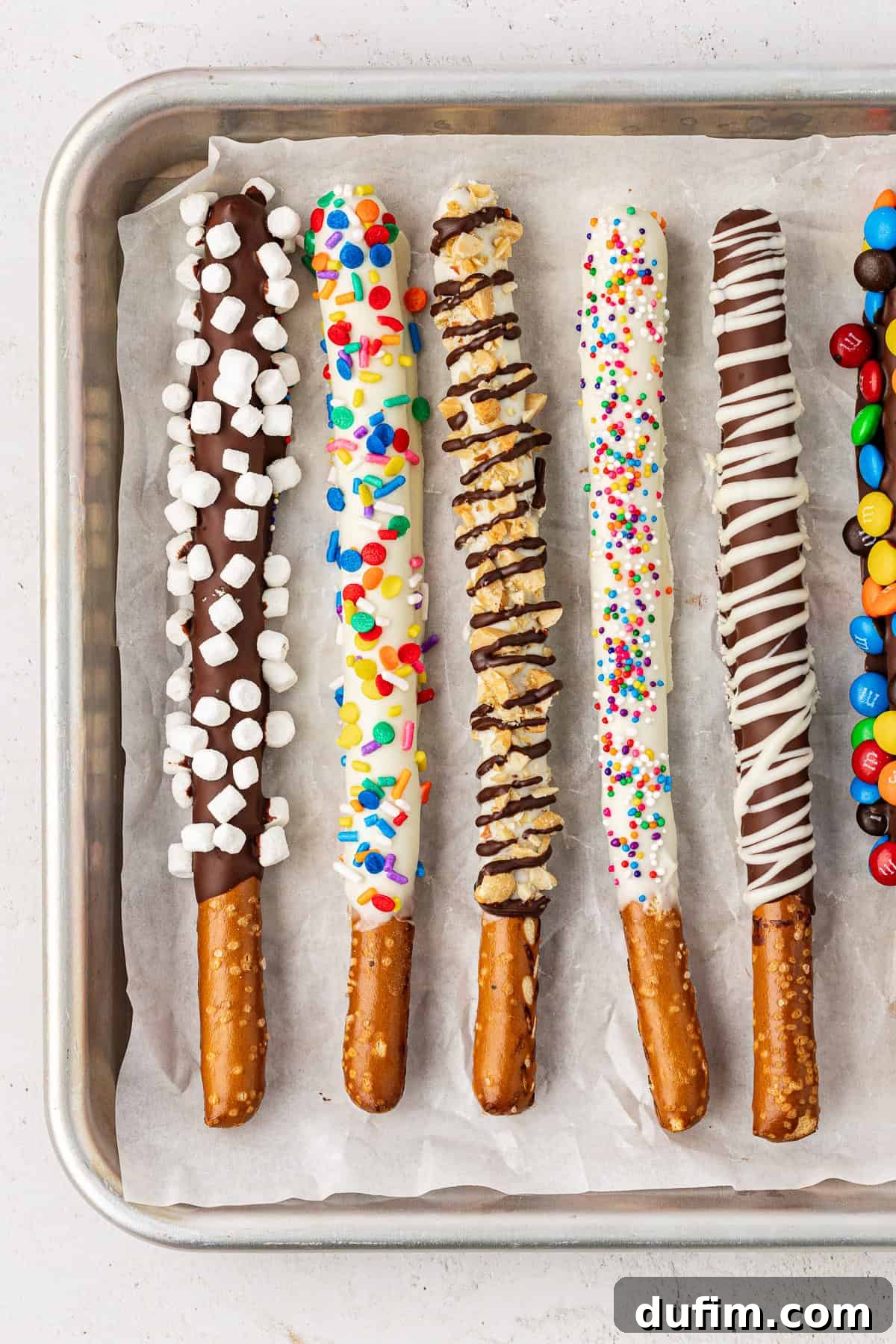 A visually appealing row of chocolate covered pretzel rods on a baking sheet, featuring various toppings: dark chocolate with mini marshmallows, white chocolate with rainbow sprinkles, white chocolate with crushed nuts and a chocolate drizzle, white chocolate with mini rainbow sprinkles, dark chocolate drizzled with white chocolate, and dark chocolate with M&M's.