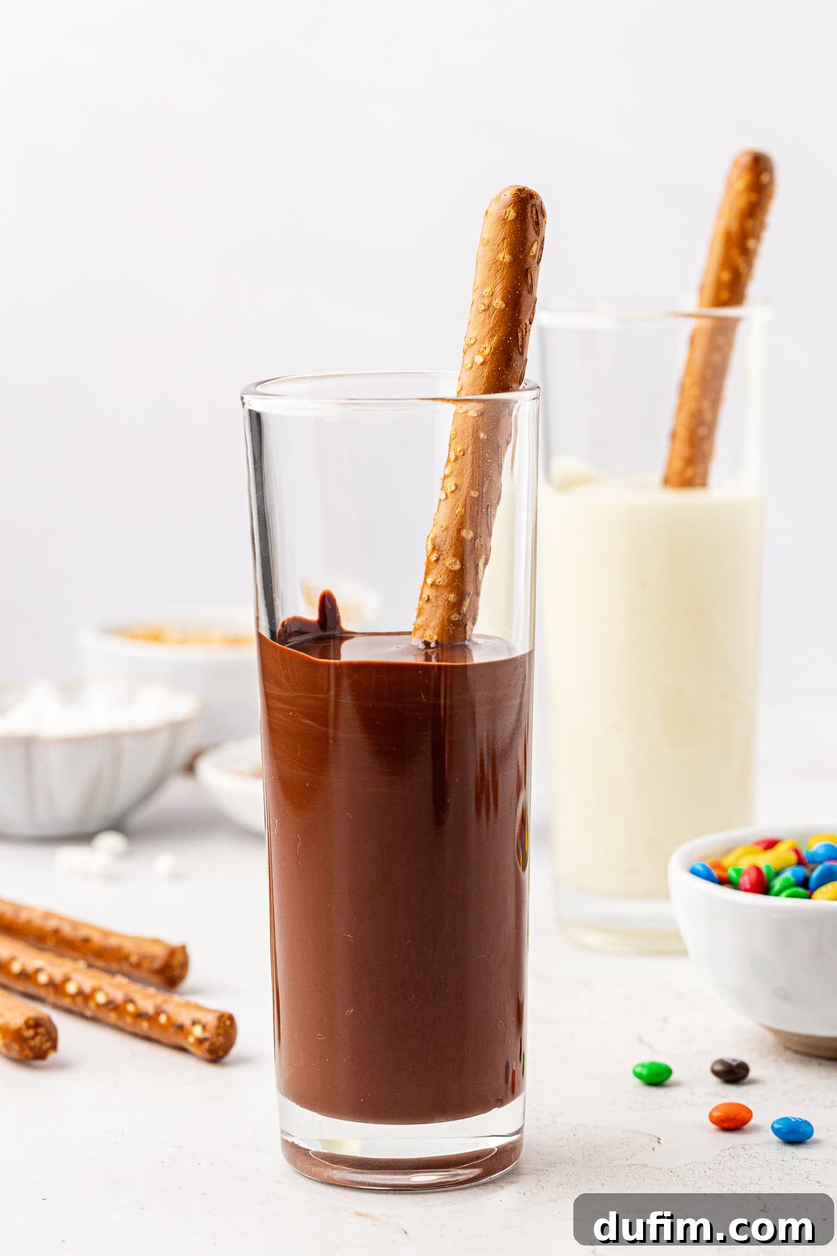 Two tall glasses, one filled with melted dark chocolate and the other with melted white chocolate, each holding a pretzel rod. Bowls of M&M's, additional pretzel rods, and more M&M's are scattered around.