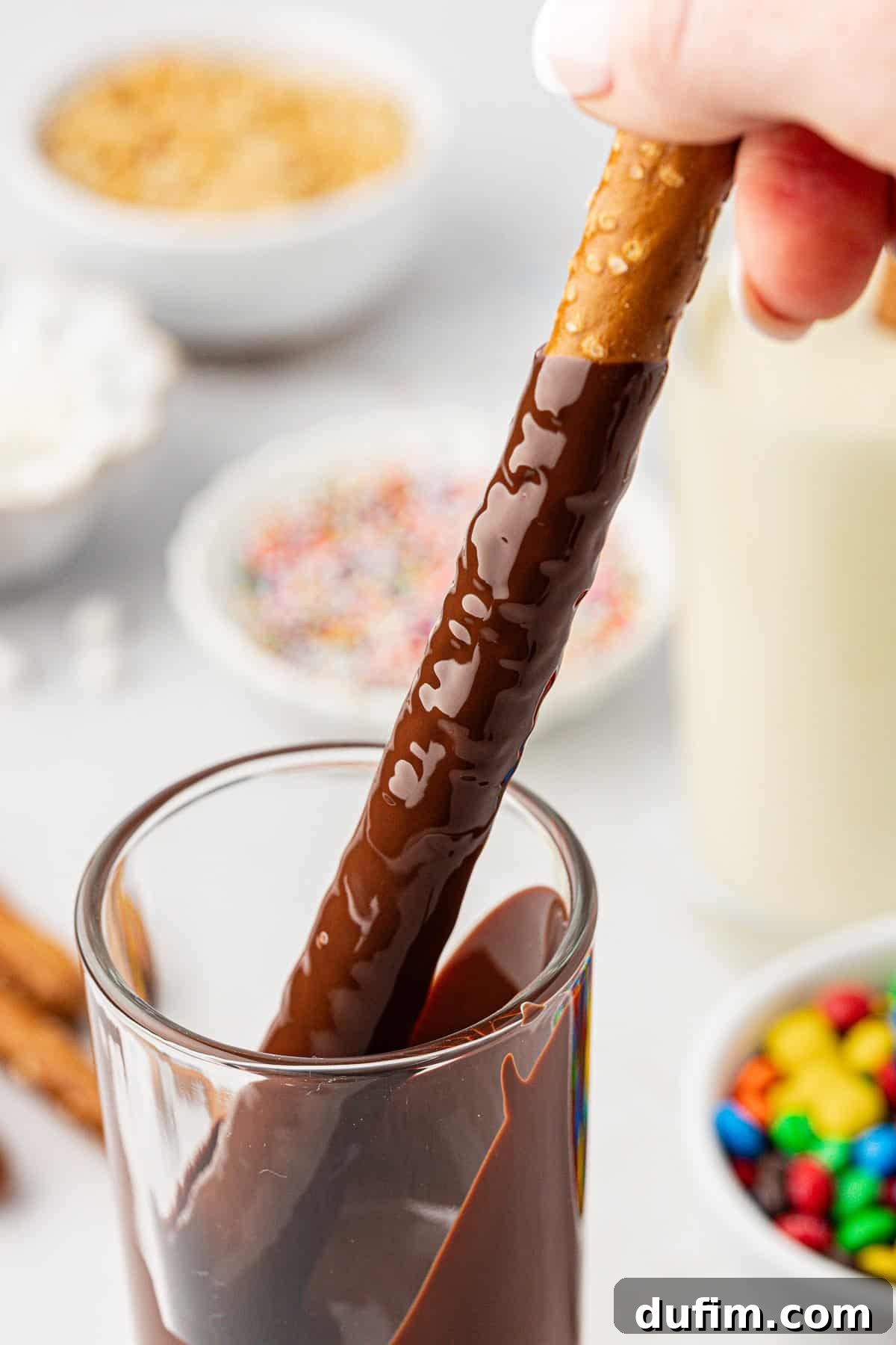 A pretzel rod being dipped into a tall glass of melted dark chocolate. In the background, several small bowls hold various toppings like M&M's and sprinkles, ready for decoration.