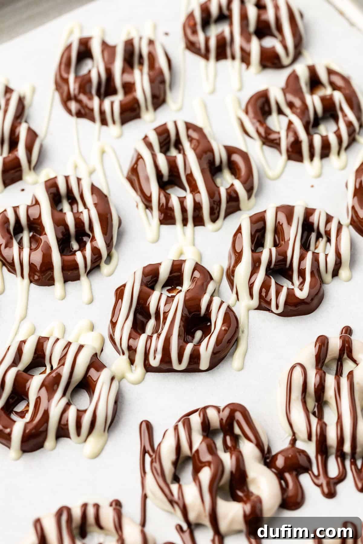 Rows of twist pretzels on a baking sheet, some coated in dark chocolate and drizzled with white chocolate, others coated in white chocolate and drizzled with dark chocolate.