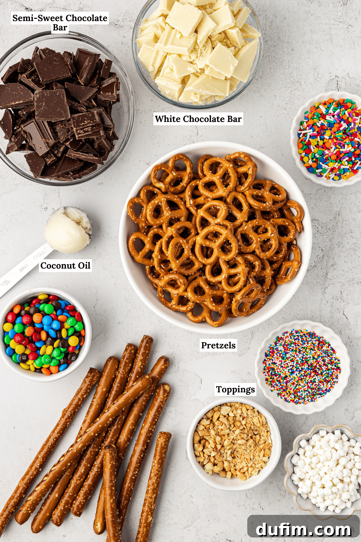 An overhead view of essential ingredients for chocolate covered pretzels: semi-sweet chocolate bars, white chocolate bars, coconut oil, vibrant rainbow sprinkles, M&M's, crushed nuts, mini marshmallows, twist pretzels, and pretzel rods.
