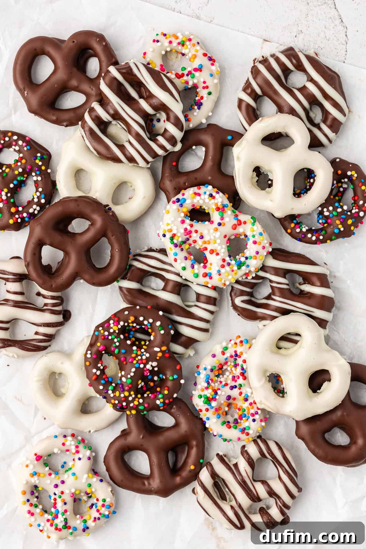 An artfully arranged collection of chocolate covered pretzels, showcasing varieties like dark chocolate with white chocolate drizzle, white chocolate with dark chocolate drizzle, and both dark and white chocolate coated in colorful rainbow sprinkles.
