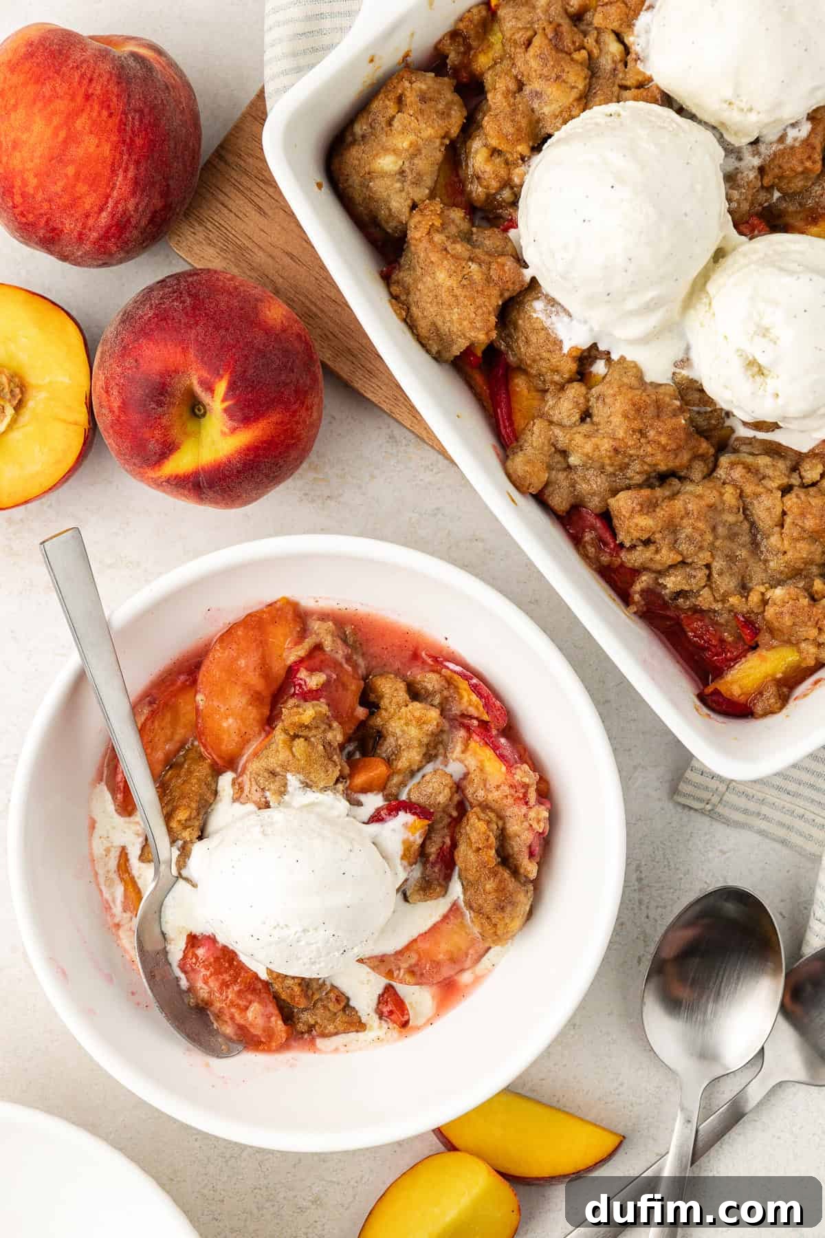 Irresistible Homemade Peach Cobbler 2 Overhead view of a bowl of peach cobbler with a scoop of vanilla ice cream and a spoon, alongside the baking dish of cobbler, fresh whole and sliced peaches, and two spoons.