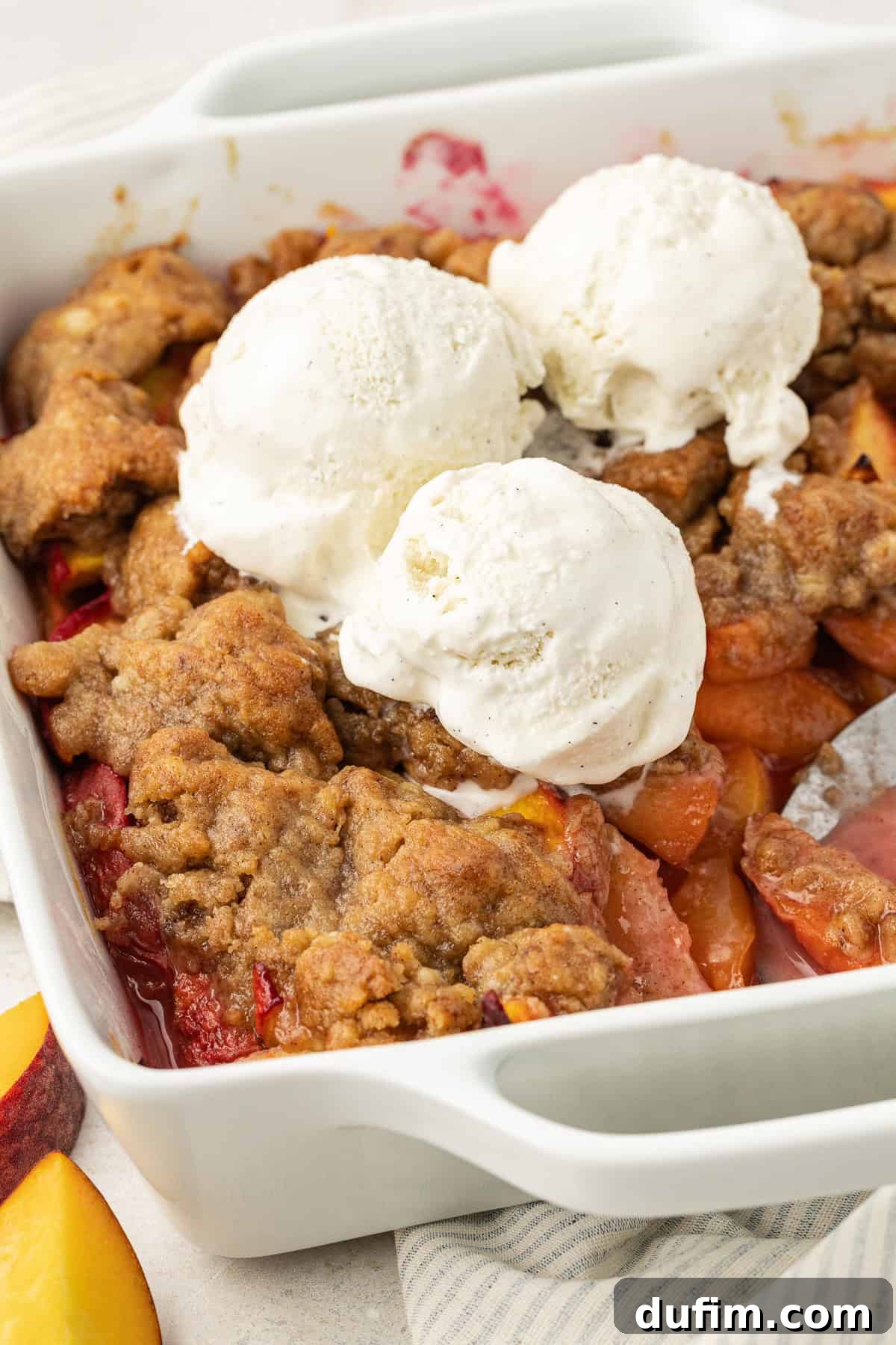 Irresistible Homemade Peach Cobbler 11 Peach cobbler in a square white baking dish, generously topped with three scoops of creamy vanilla ice cream, ready to be served.