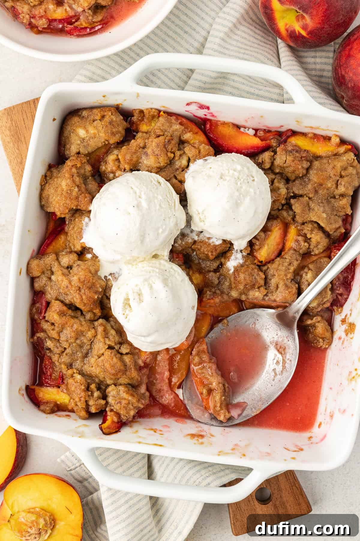 Irresistible Homemade Peach Cobbler 12 Overhead view of a baking dish of peach cobbler with three scoops of vanilla ice cream on top, a portion scooped out with a spoon left in the dish, surrounded by fresh peach slices.