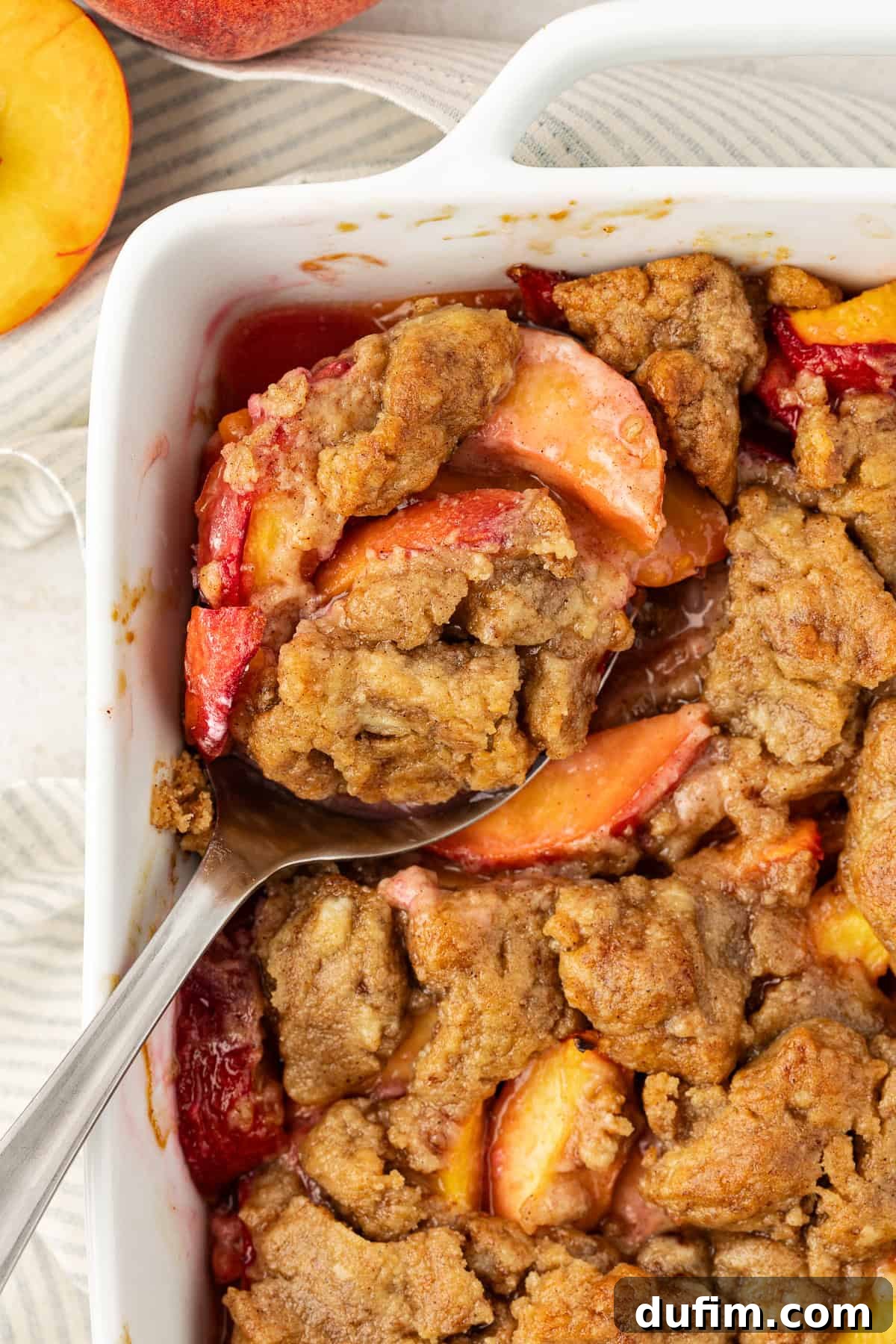 Irresistible Homemade Peach Cobbler 13 Close-up of baked peach cobbler in a white baking dish with a spoon resting in it, and fresh whole peaches to the left of the dish.