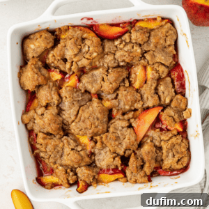 Overhead view of peach cobbler in a square white baking dish surrounded by fresh peaches, whole and slices, a stack of plates and spoon and a white and grey striped kitchen towel