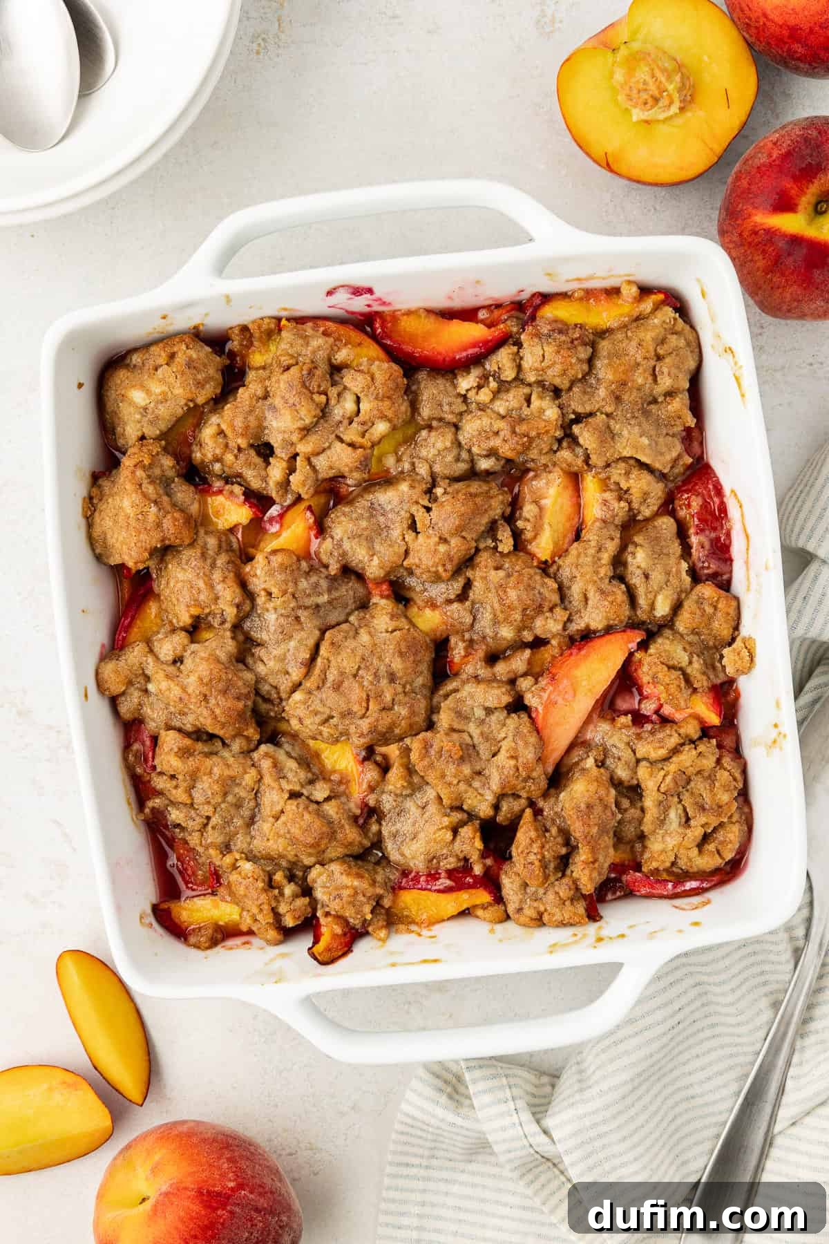 Irresistible Homemade Peach Cobbler 3 Overhead view of peach cobbler in a square white baking dish, surrounded by fresh whole and sliced peaches, a stack of plates, a spoon, and a white and gray striped kitchen towel.