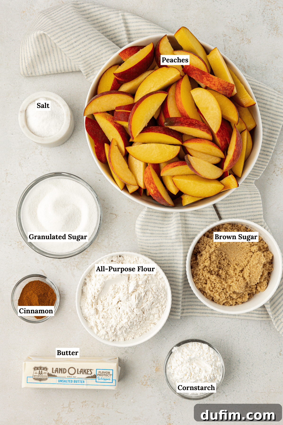 Irresistible Homemade Peach Cobbler 4 Overhead view of the organized ingredients for peach cobbler, including sliced peaches, salt, granulated sugar, brown sugar, all-purpose flour, cinnamon, butter, and cornstarch, laid out on a white surface.