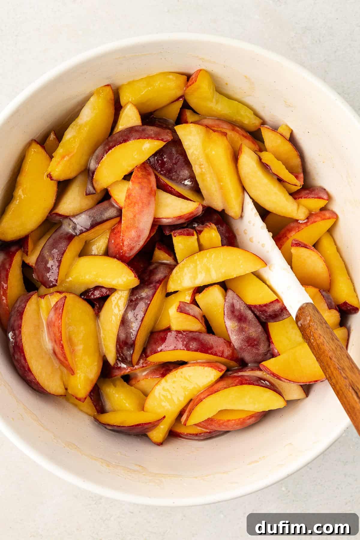 Irresistible Homemade Peach Cobbler 5 Fresh peach slices being gently mixed with sugar and cornstarch in a white bowl using a white spatula, preparing the peach filling for the cobbler.