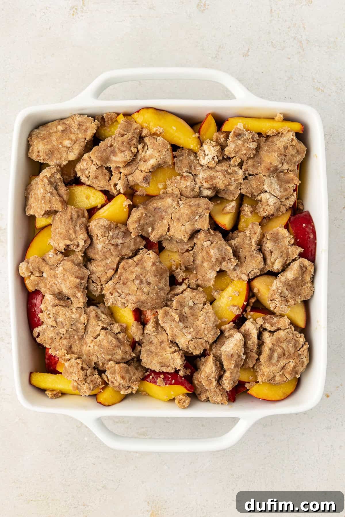 Irresistible Homemade Peach Cobbler 8 Peach cobbler assembled and ready to be baked in a square white baking dish, showing the peach filling topped with the crumble mixture.