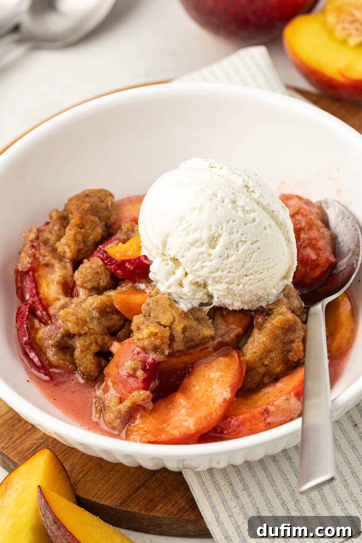 Irresistible Homemade Peach Cobbler 10 A single serving of peach cobbler in a white bowl, topped with a scoop of vanilla ice cream and a spoon, resting on a wooden platter with a white and gray striped kitchen towel. Fresh peach slices are scattered around.