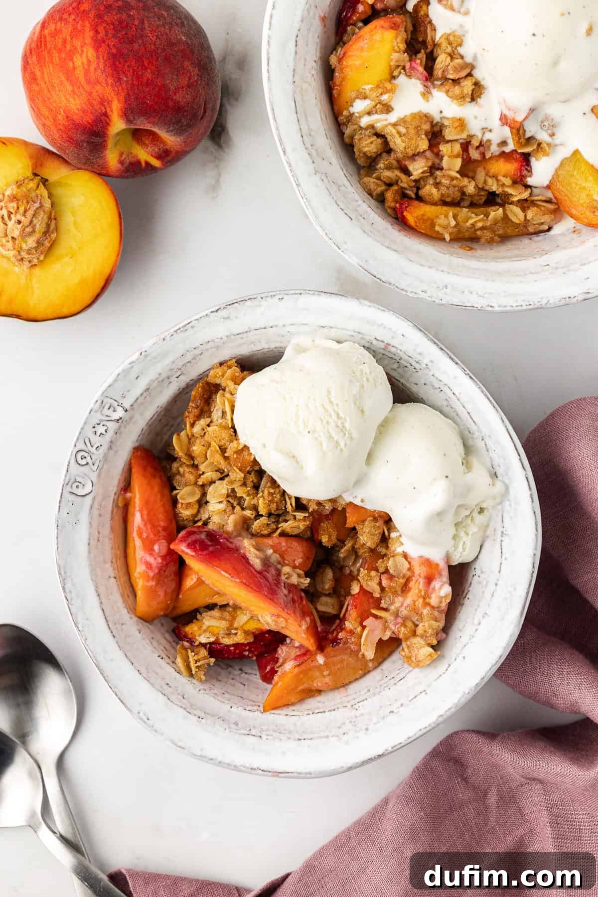 Two white bowls of peach crisp, each topped with a generous scoop of vanilla ice cream. The bowls are set on a light purple kitchen towel, accompanied by two spoons and fresh peaches, creating an inviting summer dessert scene.