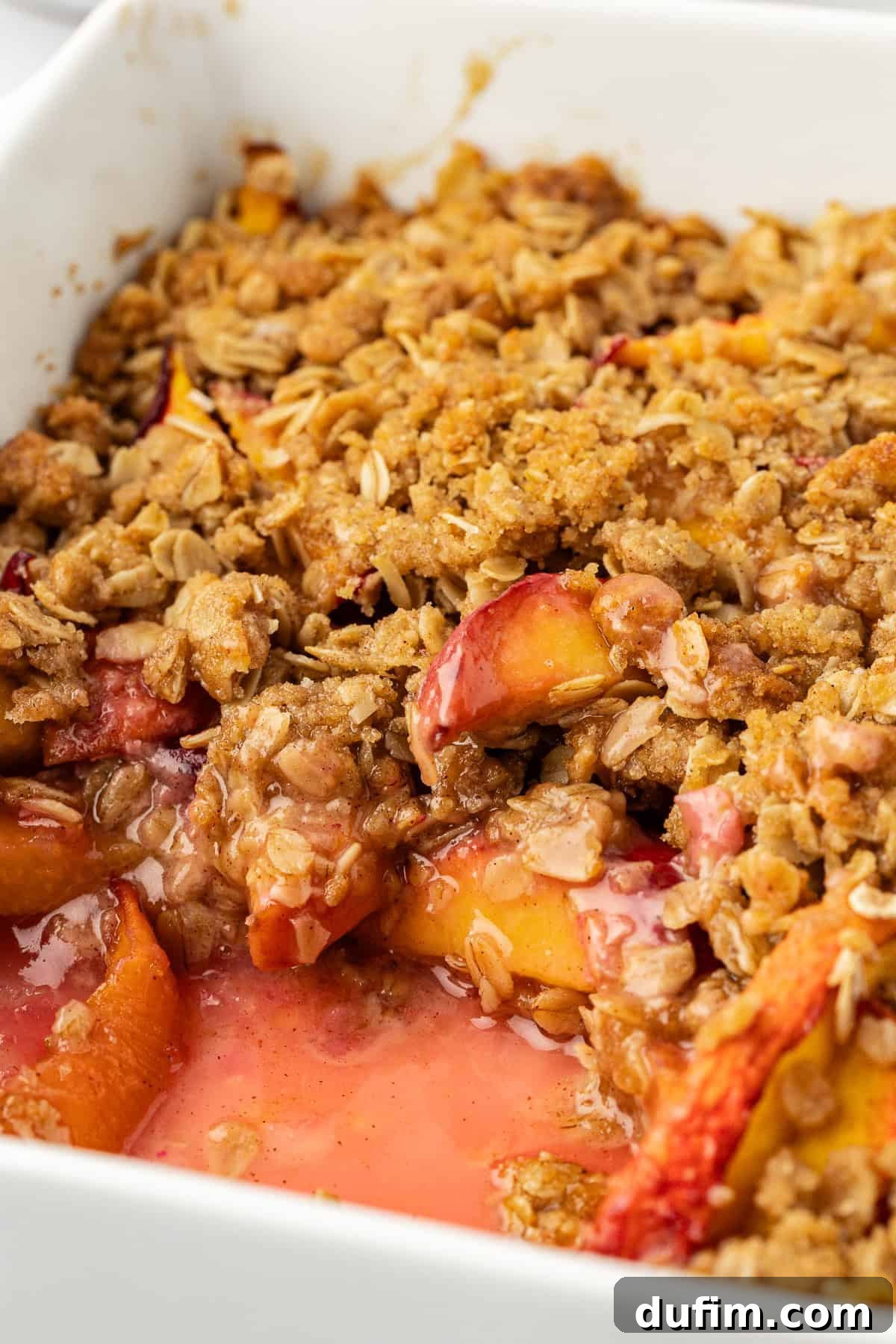 Close-up view of a baked peach crisp in a white baking dish, with a scoop already removed, revealing the golden-brown, bubbly fruit filling beneath the crispy topping.