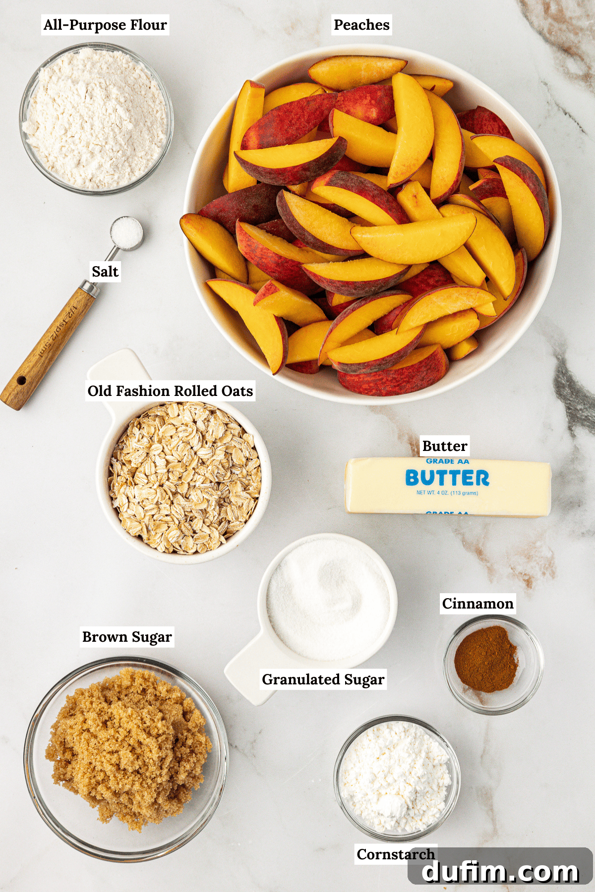 Overhead view of all the ingredients for peach crisp laid out on a white surface: sliced fresh peaches, old-fashioned rolled oats, all-purpose flour, brown sugar, granulated sugar, cinnamon, salt, cold unsalted butter, and cornstarch.