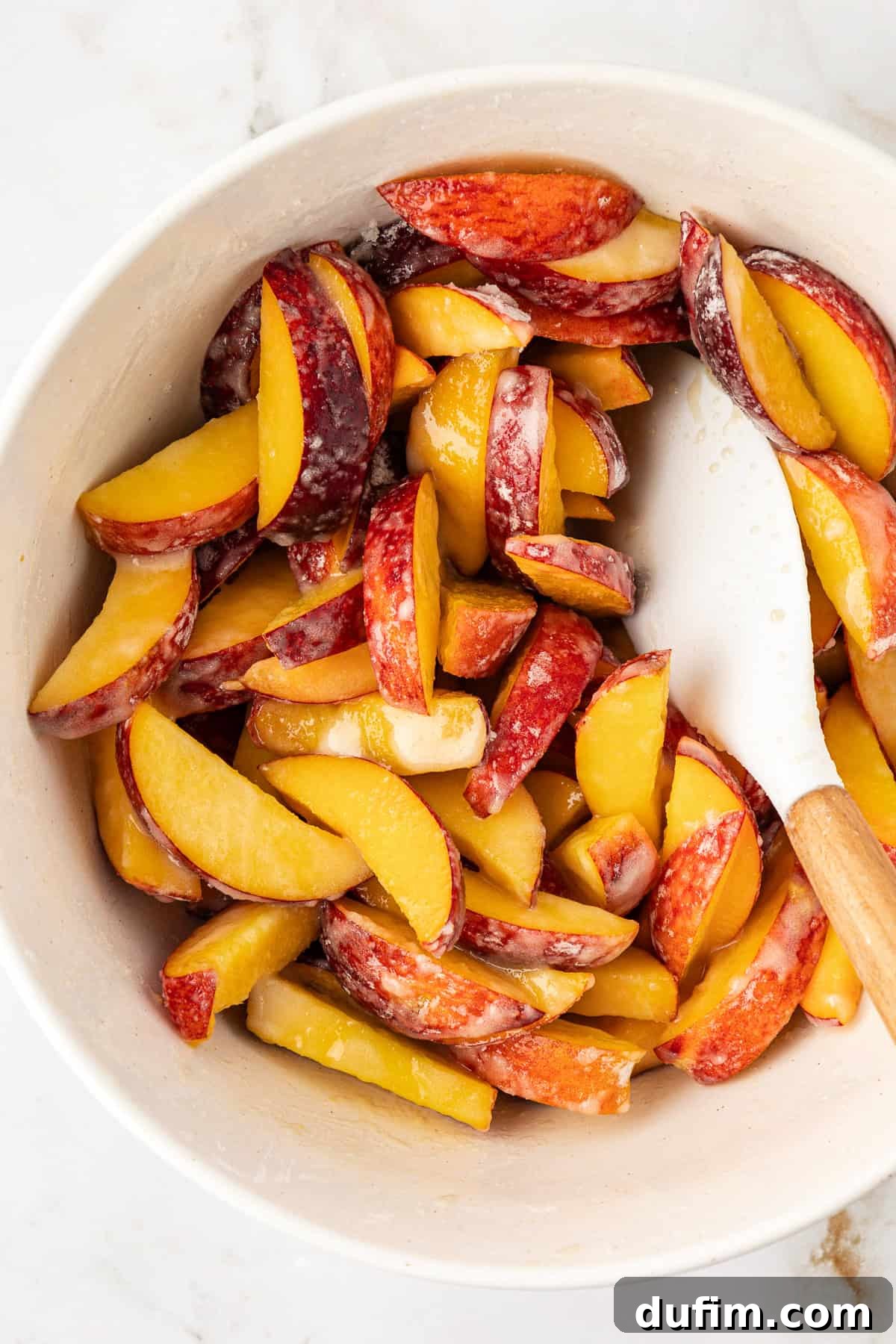 A bowl of freshly sliced peaches being gently mixed with a white and wooden spatula, ready for the peach filling preparation.
