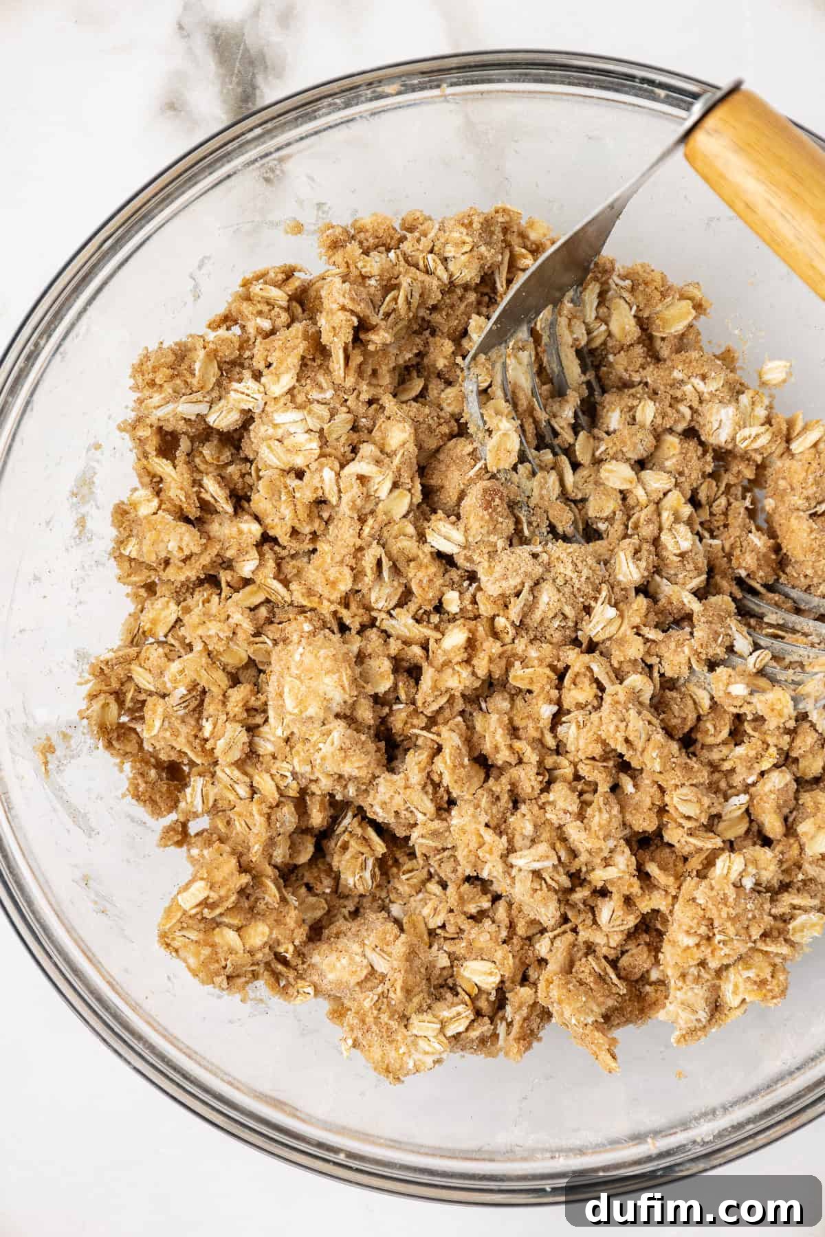 A clear glass mixing bowl filled with the crumbly peach crisp topping mixture, showing the oats, flour, sugars, and small pieces of butter.