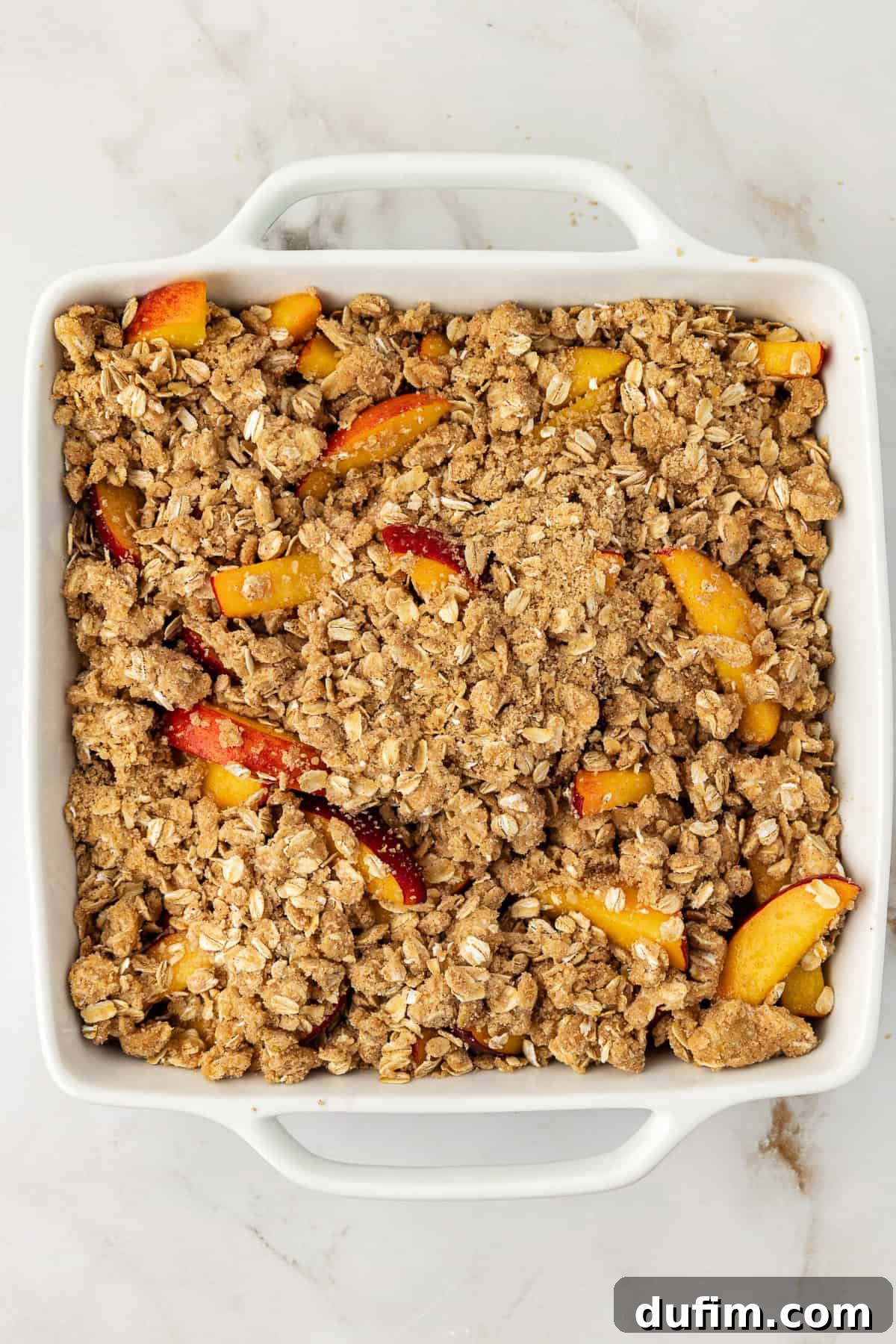 Overhead view of a peach crisp in a square baking dish before baking, showing the even distribution of the crumbly topping over the sliced peaches.