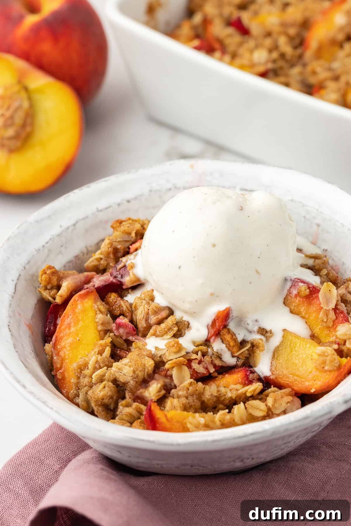 A small white bowl containing a serving of peach crisp, topped with a scoop of melting vanilla ice cream, resting on a light purple kitchen towel. In the background, fresh peaches and the larger baking dish of peach crisp are visible, highlighting the deliciousness of the dessert.