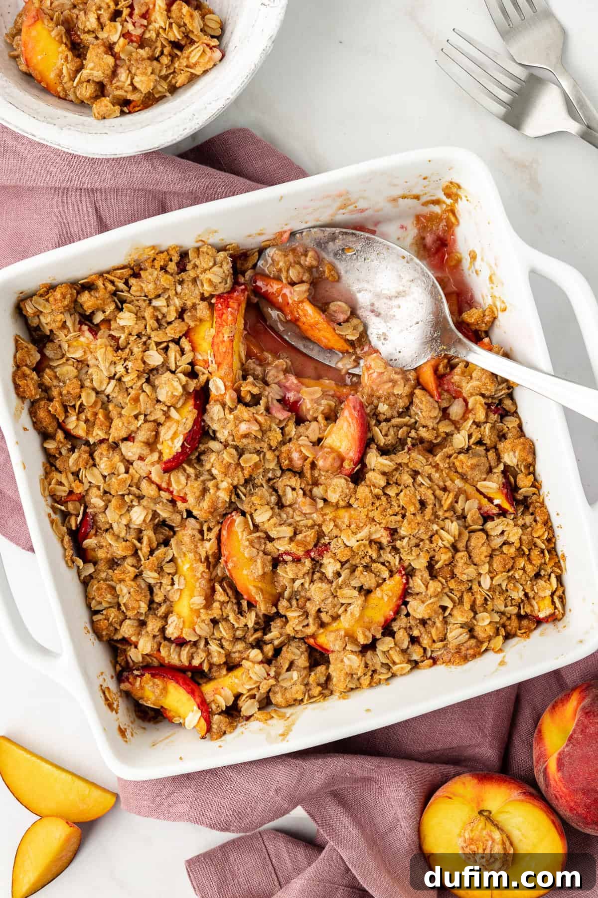 Overhead view of a baking dish of peach crisp with a serving spoon, and a scoop already missing. It sits on a light purple kitchen towel, surrounded by a smaller bowl of peach crisp, forks, and both sliced and whole fresh peaches, creating a bountiful dessert spread.