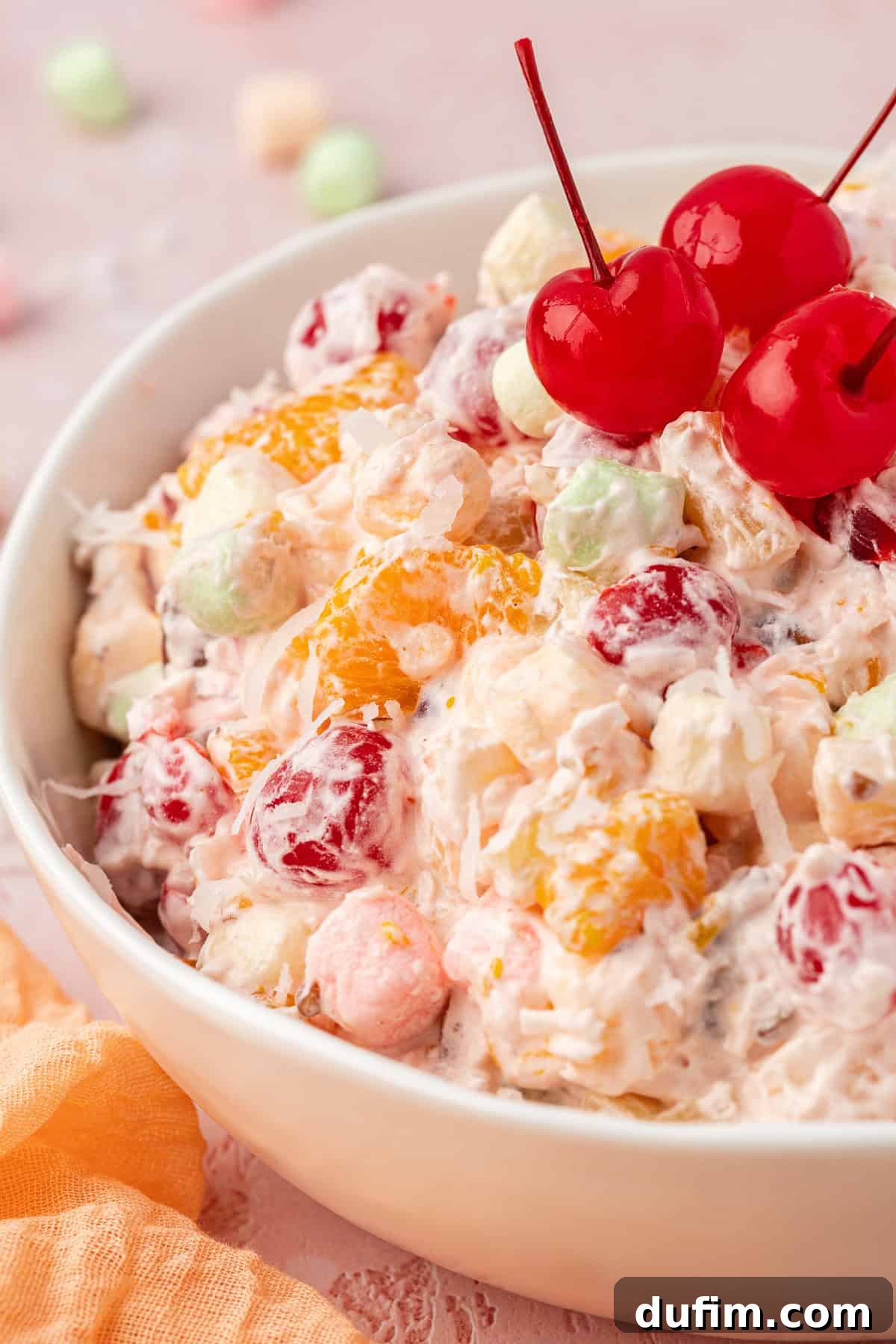 Grandma's Favorite Ambrosia Salad 2 Close-up of a white bowl filled with creamy Ambrosia Salad, generously garnished with three bright red maraschino cherries. A light orange towel rests invitingly beside the bowl, with a scattering of miniature marshmallows around, hinting at the dish's delightful components.