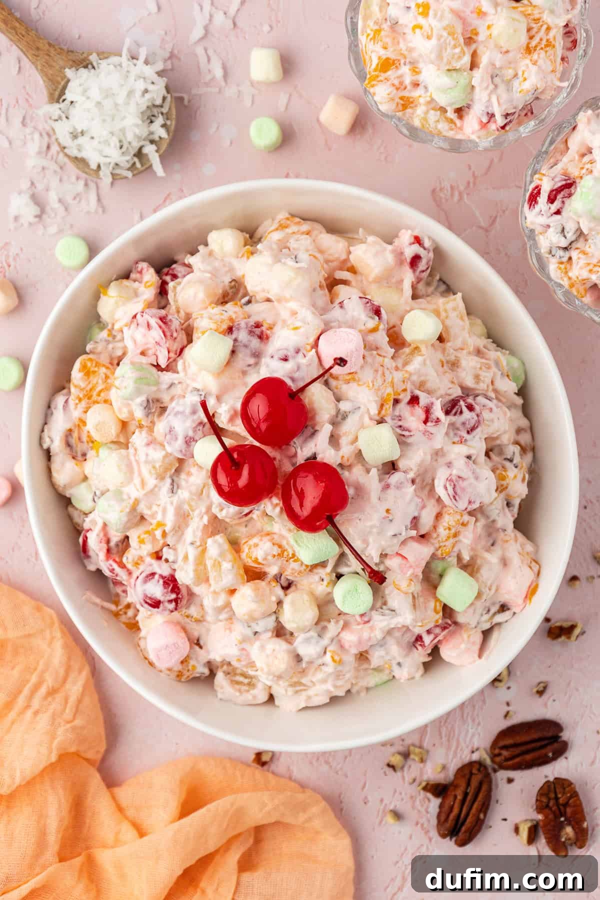 Grandma's Favorite Ambrosia Salad 3 Overhead view of a large white bowl brimming with perfectly prepared Ambrosia Salad, elegantly topped with three vibrant cherries. The appealing scene is completed with two smaller dishes of ambrosia, a spoon holding delicate coconut flakes, a soft light orange towel, and miniature marshmallows and pecans artfully scattered around, highlighting the salad's delightful ingredients.