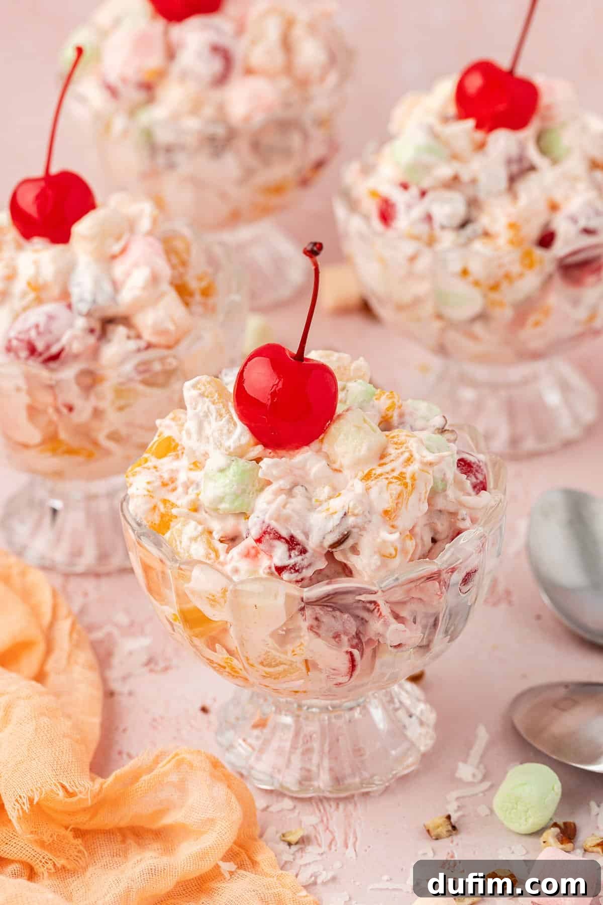 Grandma's Favorite Ambrosia Salad 4 Four small, elegant trifle dishes filled with Ambrosia Salad, each topped with a glistening red cherry. They are artfully arranged beside a soft light orange towel and shiny spoons, with miniature marshmallows, shredded coconut, and small pecan pieces scattered charmingly around, suggesting a festive and delicious serving.