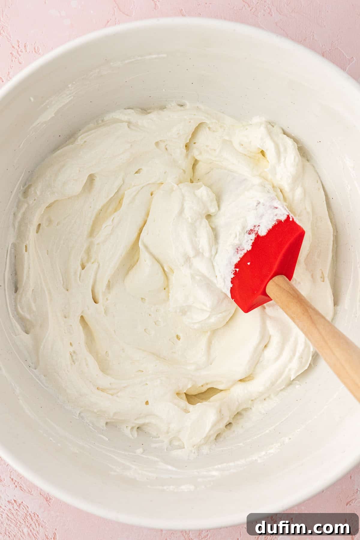 Grandma's Favorite Ambrosia Salad 6 Sour cream and whipped topping being gently mixed together with a spatula in a pristine white bowl, resting on a soft pink surface, illustrating the first step of combining the creamy base ingredients to create a smooth and uniform mixture.