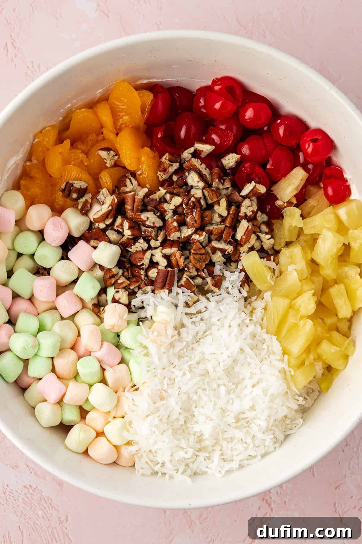 Grandma's Favorite Ambrosia Salad 7 A vibrant array of Ambrosia Salad ingredients unmixed in a large white bowl: colorful mini marshmallows, bright mandarin oranges, halved maraschino cherries, chopped pecans, sweet pineapple tidbits, and fluffy coconut flakes, all waiting to be gently folded into the creamy base, showcasing the rich variety of components.