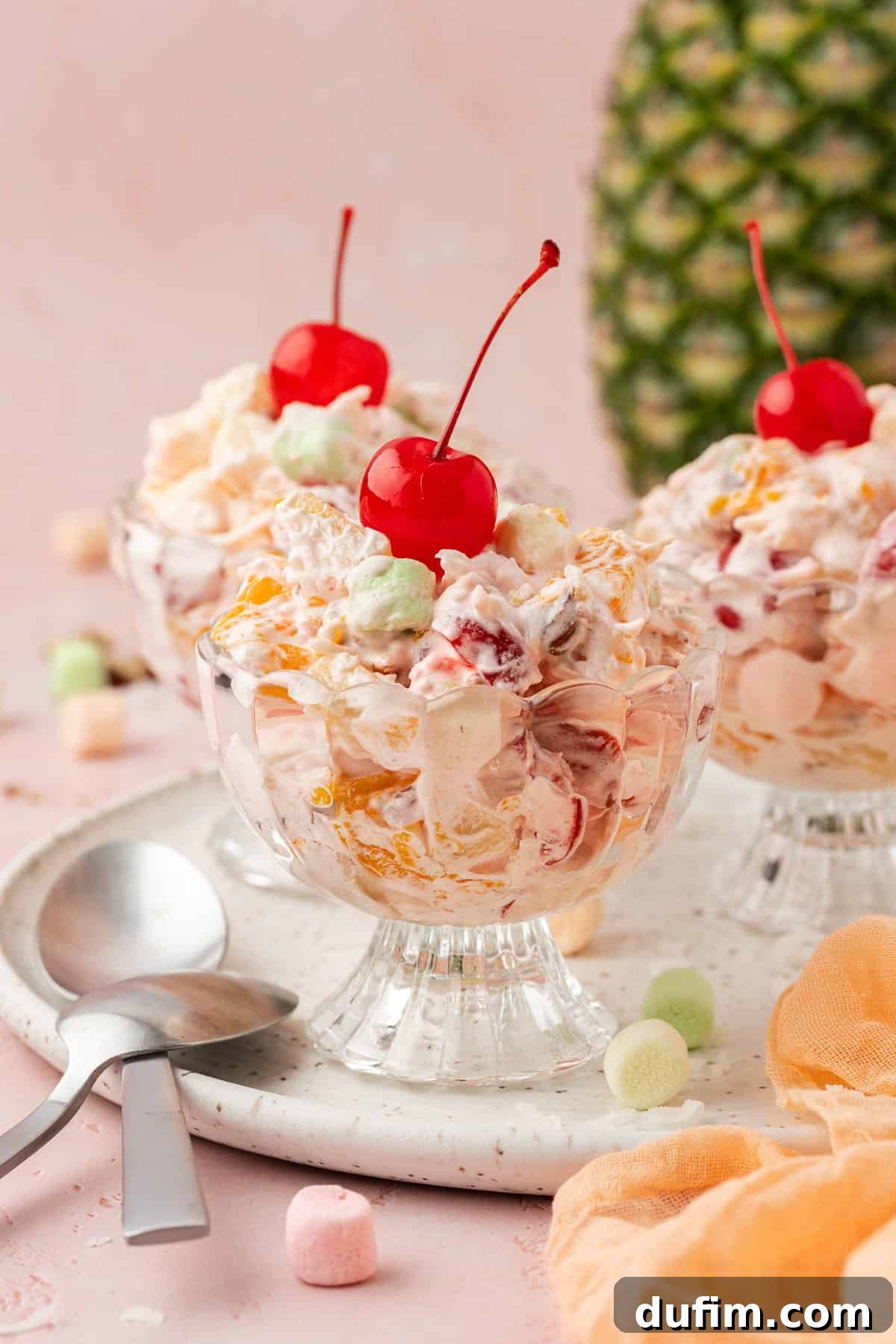 Grandma's Favorite Ambrosia Salad 8 Three small, elegant trifle dishes brimming with perfectly prepared Ambrosia Salad, artfully arranged on a pristine white platter with spoons resting beside them. A whole pineapple stands in the softly blurred background, complemented by a light orange towel and scattered mini marshmallows, suggesting a delightful and ready-to-serve dessert.