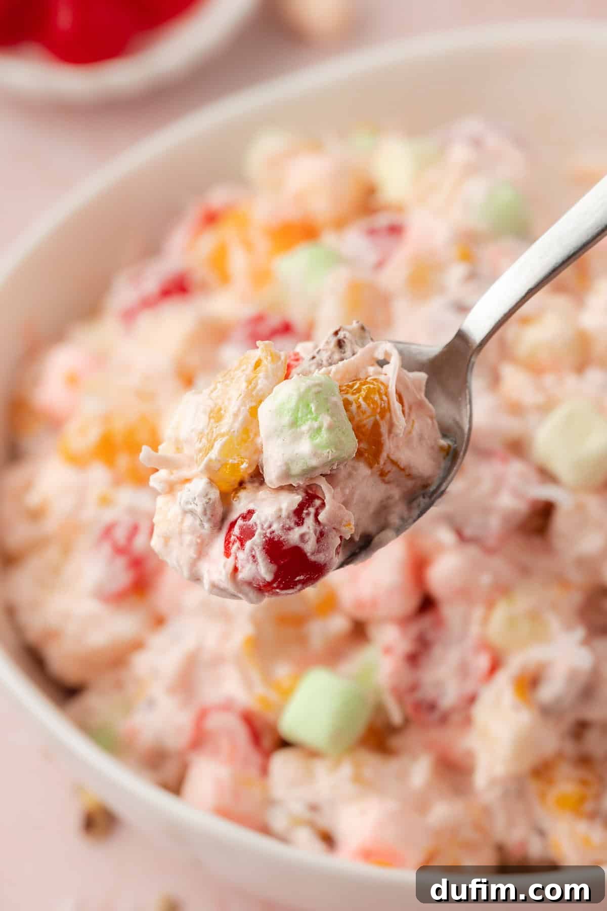 Grandma's Favorite Ambrosia Salad 9 A spoonful of creamy Ambrosia Salad, adorned with colorful fruit and fluffy marshmallows, is being gently lifted from a large bowl, showcasing its delightful texture and inviting, vibrant appearance, ready to be enjoyed.