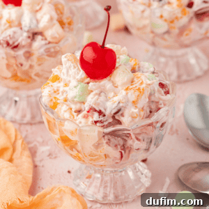 A small trifle dish filled with beautifully crafted Ambrosia Salad, crowned with a vibrant maraschino cherry, surrounded by more servings of the salad, a soft light orange towel, and a spoon, inviting you to indulge in its creamy, fruity goodness.