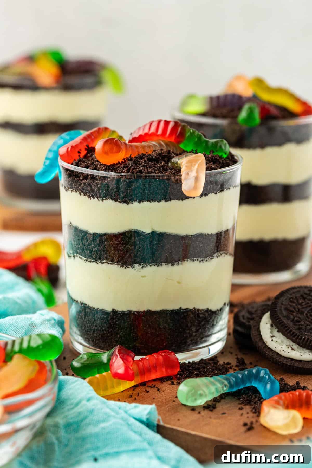 Mastering Delicious Dirt Cake 14 Three small individual trifle dishes of dirt cake, each artfully topped with playful gummy worms, arranged on a rustic wooden platter. The display is complemented by whole Oreo cookies, crushed Oreo crumbs, extra gummy worms, and a soft light teal towel, creating an inviting and fun dessert presentation.