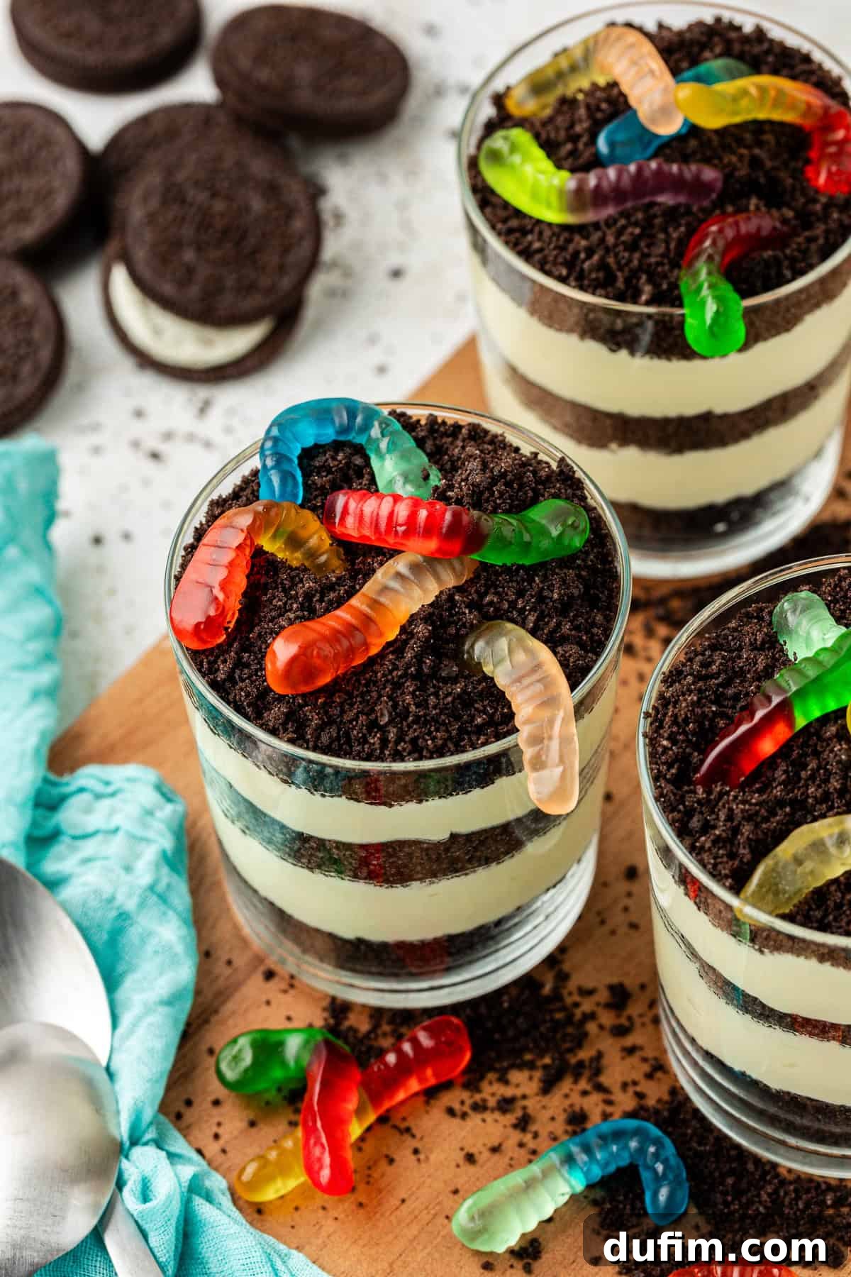 Mastering Delicious Dirt Cake 15 A trio of charming individual trifle dishes filled with rich dirt cake, elegantly topped with playful gummy worms, arranged on a rustic wooden platter. Surrounding the dishes are whole Oreo cookies, fine Oreo crumbs, extra gummy worms, and spoons, all resting on a soft light teal towel, creating a beautiful and appetizing dessert spread.