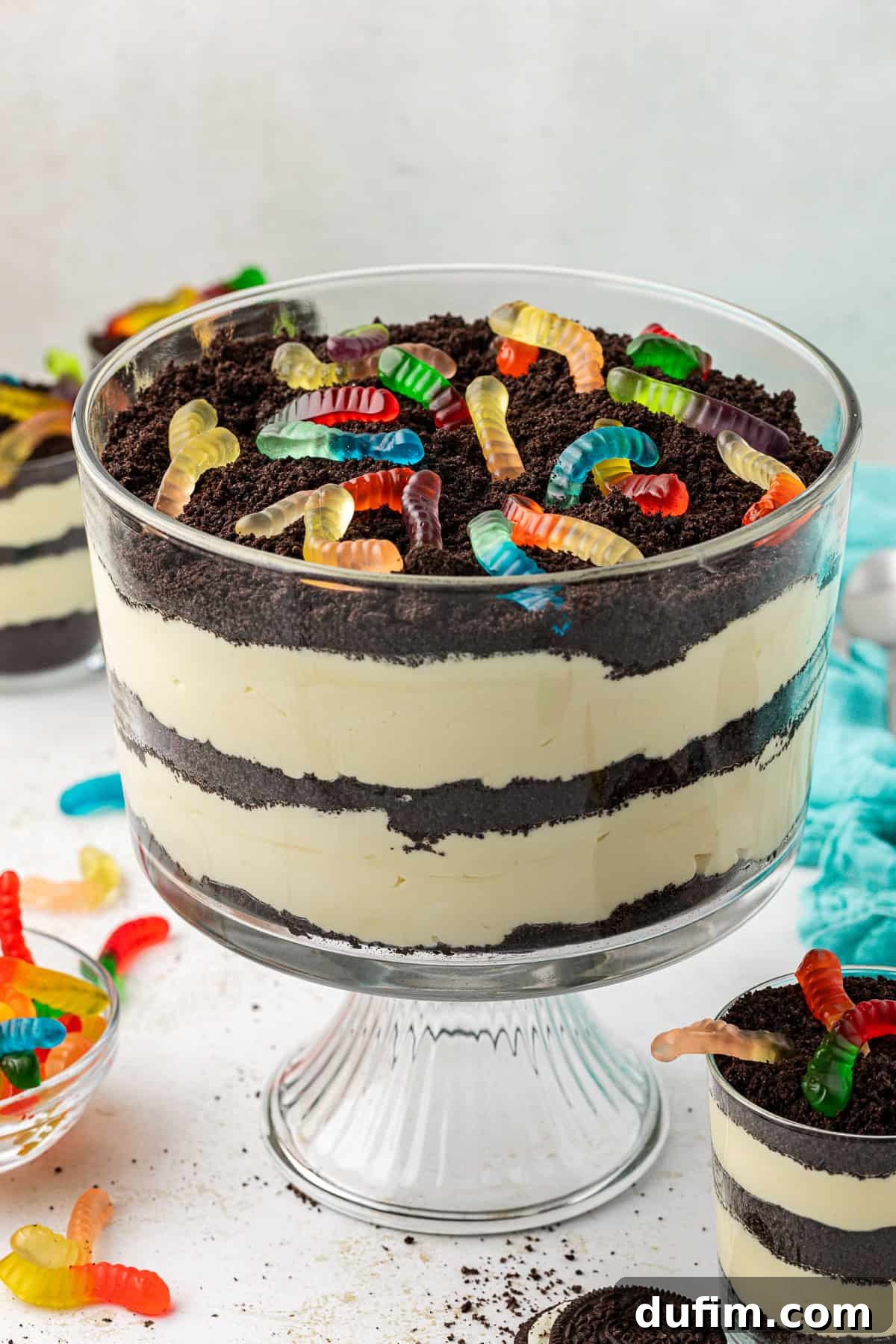 Mastering Delicious Dirt Cake 3 A large clear glass trifle dish brimming with homemade dirt cake and whimsical gummy worms, surrounded by smaller individual dirt cake trifles, a soft teal towel, extra gummy worms, and whole Oreo cookies, ready for a fun party.