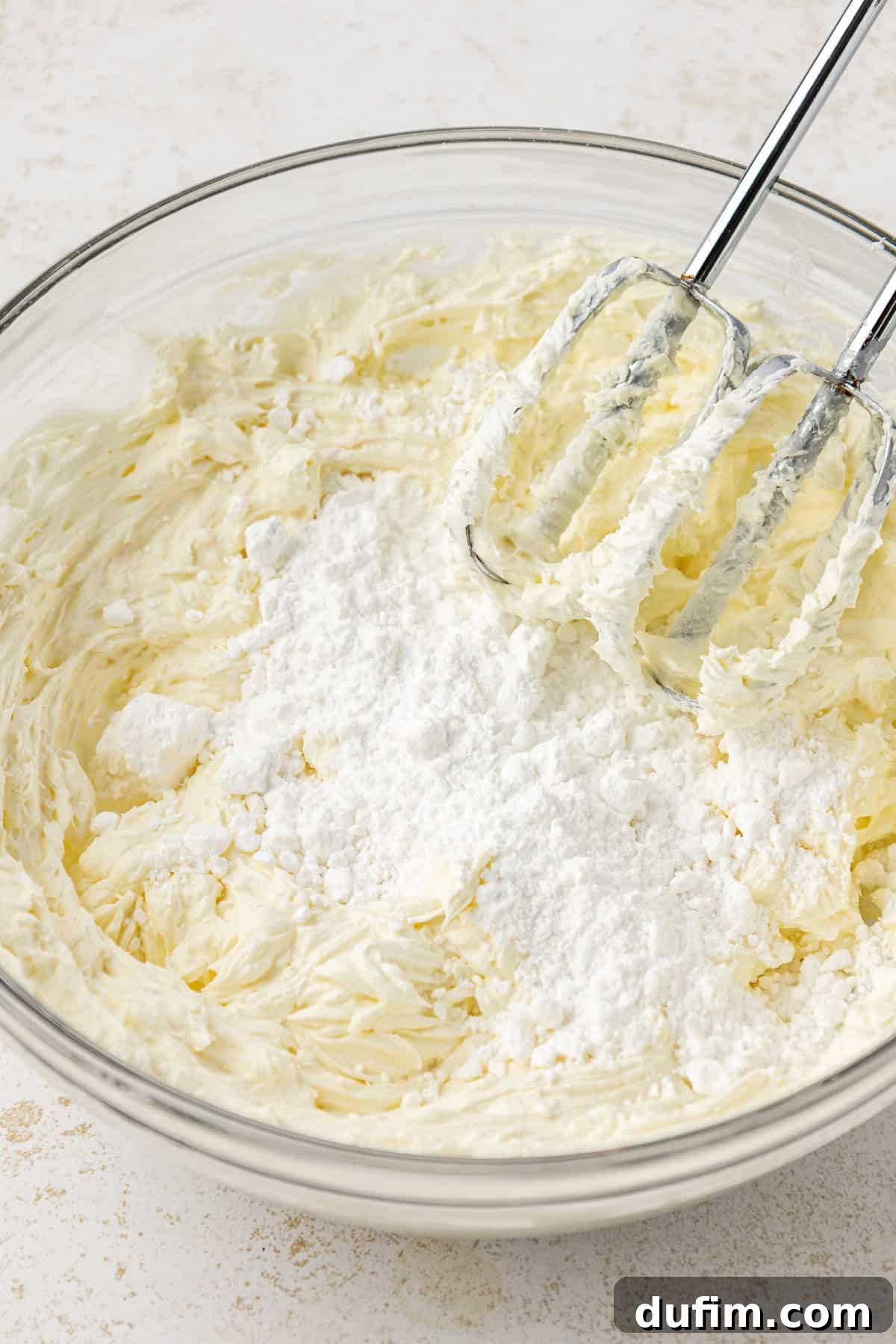 Mastering Delicious Dirt Cake 7 A smooth mixture of cream cheese, butter, and powdered sugar being beaten to perfection in a large clear glass bowl using an electric mixer, creating a light and airy texture.