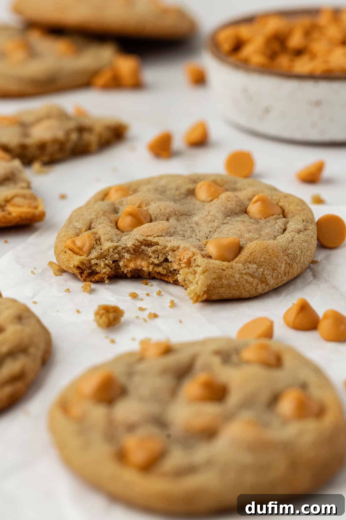 Sweet Butterscotch Chip Delights 2 Delicious butterscotch cookies scattered on white parchment paper, with one cookie bitten to show its soft, chewy interior. Cookie crumbs and extra butterscotch chips are sprinkled around.