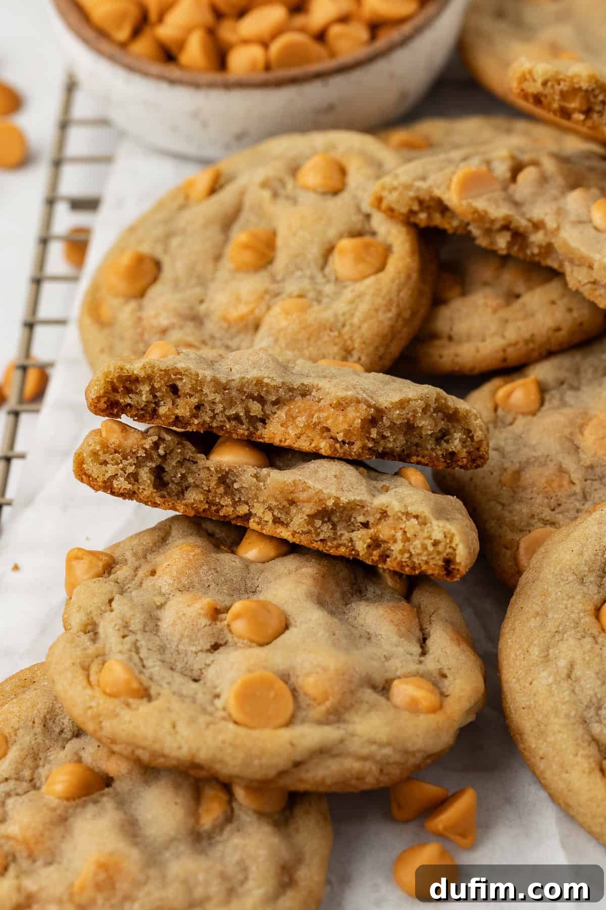 Sweet Butterscotch Chip Delights 17 Close-up view of a pile of delicious butterscotch cookies on a wire rack lined with white parchment paper. One cookie is broken in half, revealing its soft, chewy interior filled with butterscotch chips. More chips are scattered around.