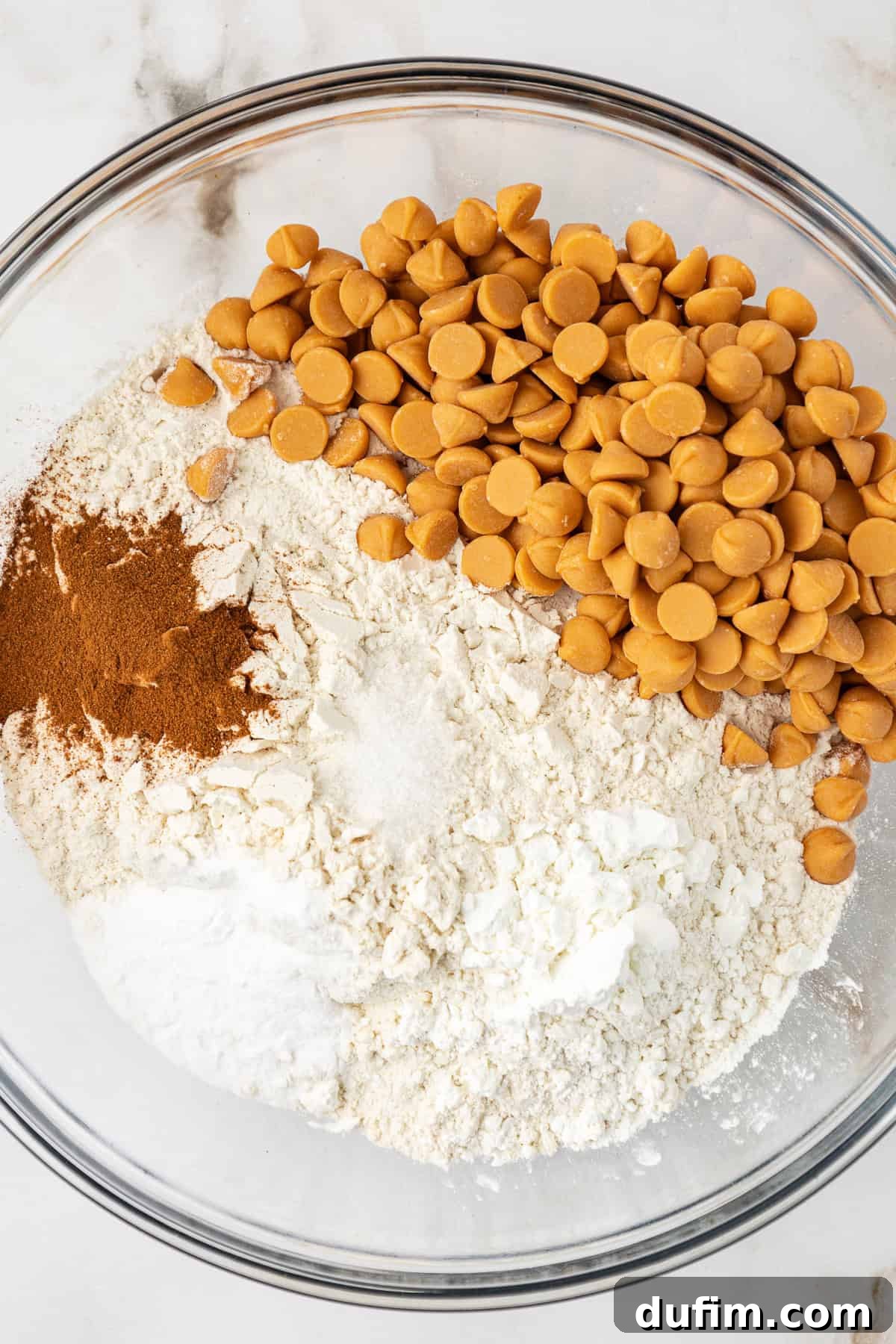 Sweet Butterscotch Chip Delights 5 Dry ingredients for butterscotch cookies in a clear glass bowl, not yet mixed, showing flour, sugar, cinnamon, cornstarch, baking soda, and butterscotch chips layered on top.