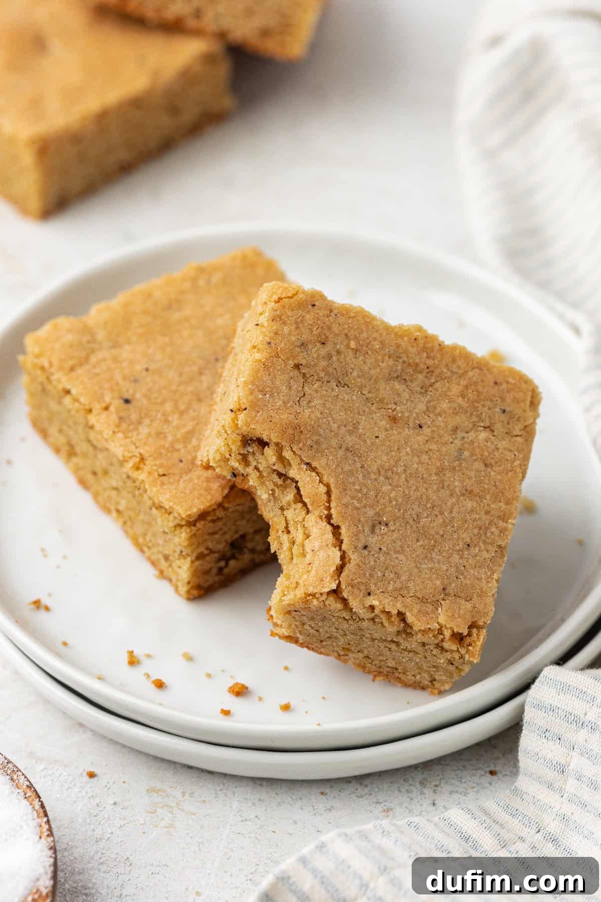 Golden Blondie Bars 4 Two golden blondie squares on stacked white plates, with one having a bite taken out, revealing its soft, tender crumb.