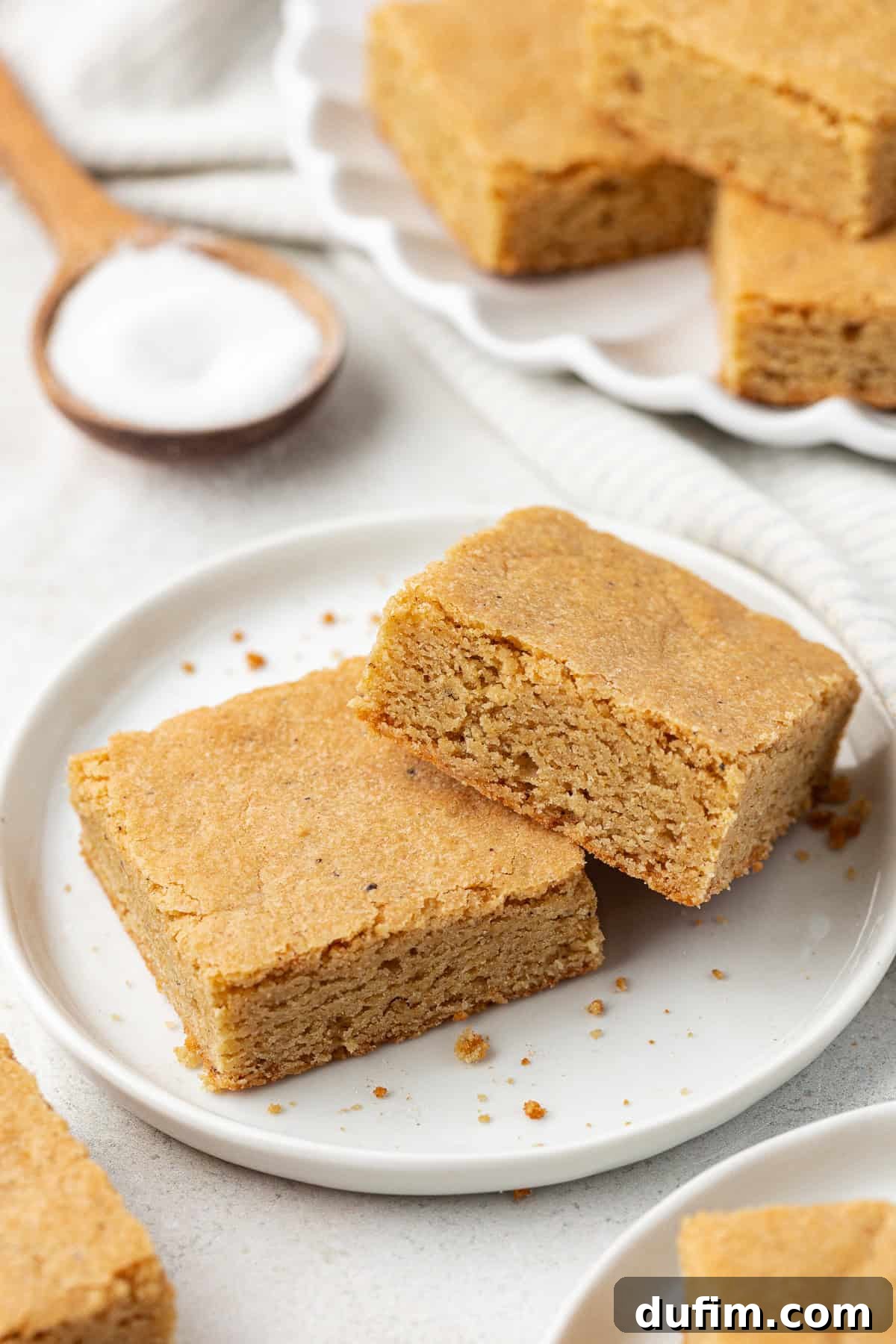 Golden Blondie Bars 5 A small white plate holds two blondie squares, surrounded by more blondies on plates and a wooden spoon brimming with brown sugar, emphasizing the key ingredient.