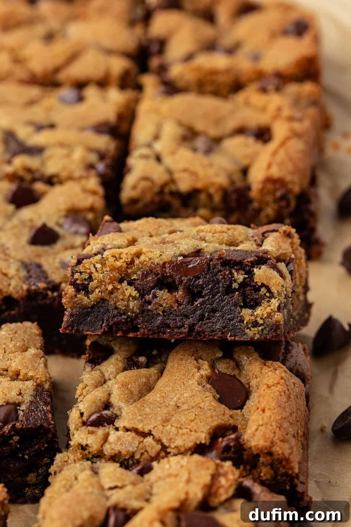 Rows of brookies on brown parchment paper, with one brookie leaning on top of another, clearly showing the two distinct layers: a rich brownie base and a golden chocolate chip cookie top, perfectly separated.