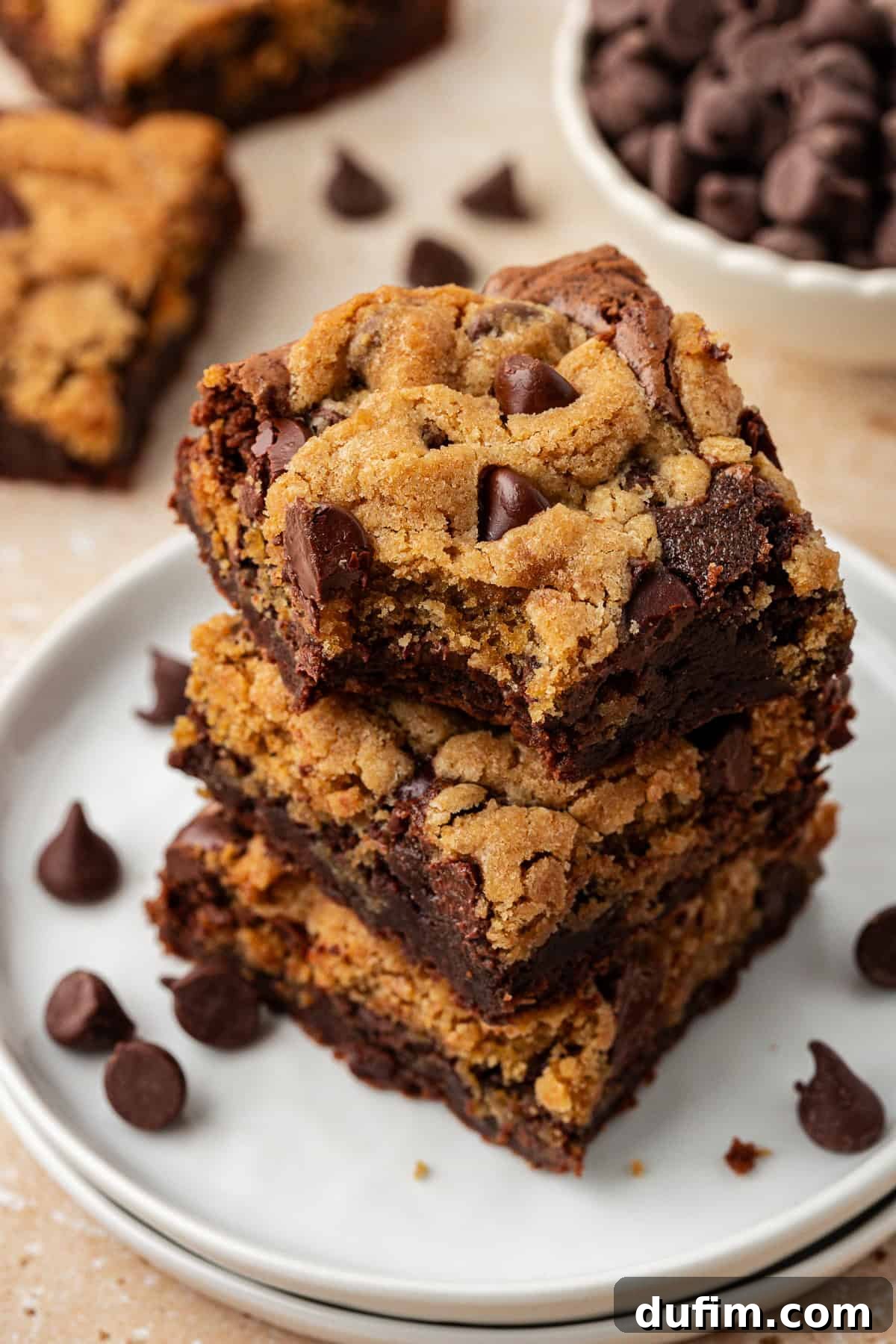 Three perfectly baked brookies stacked in a tower on a small stack of two white plates, with more brookies and loose chocolate chips artfully scattered around on a light surface, ready to be enjoyed.