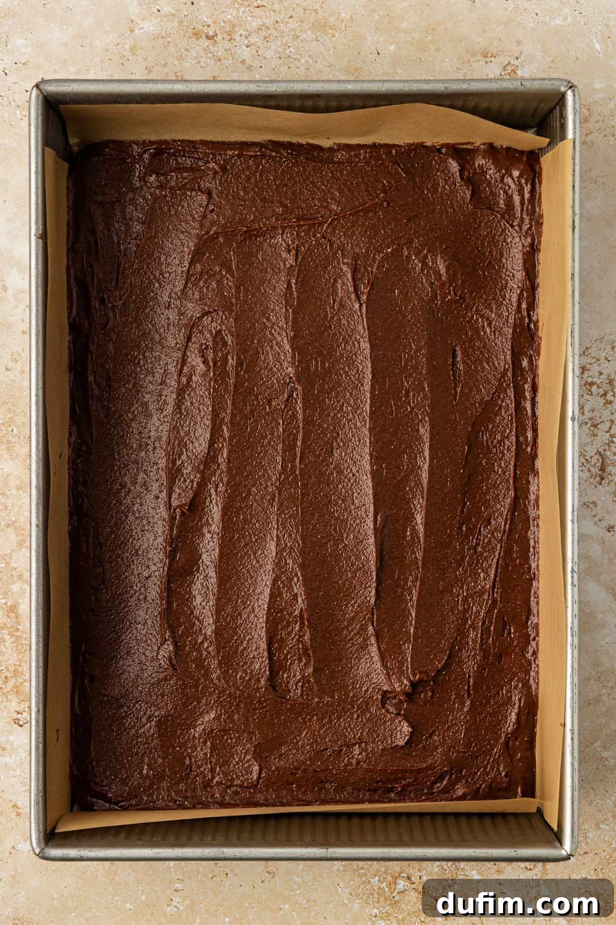 A smooth, dark brown brownie batter layer spread evenly in a metal baking pan lined with light brown parchment paper, serving as the foundational layer for the brookies.