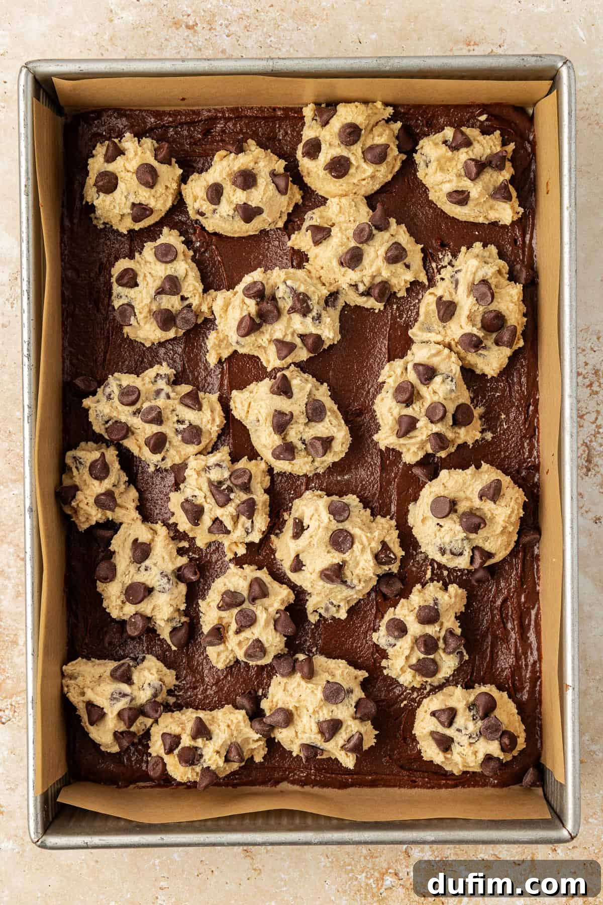 An overhead view of a metal baking pan lined with parchment paper, containing a smooth brown brownie batter base topped with evenly distributed dollops of golden chocolate chip cookie dough, creating a polka-dotted effect before baking.