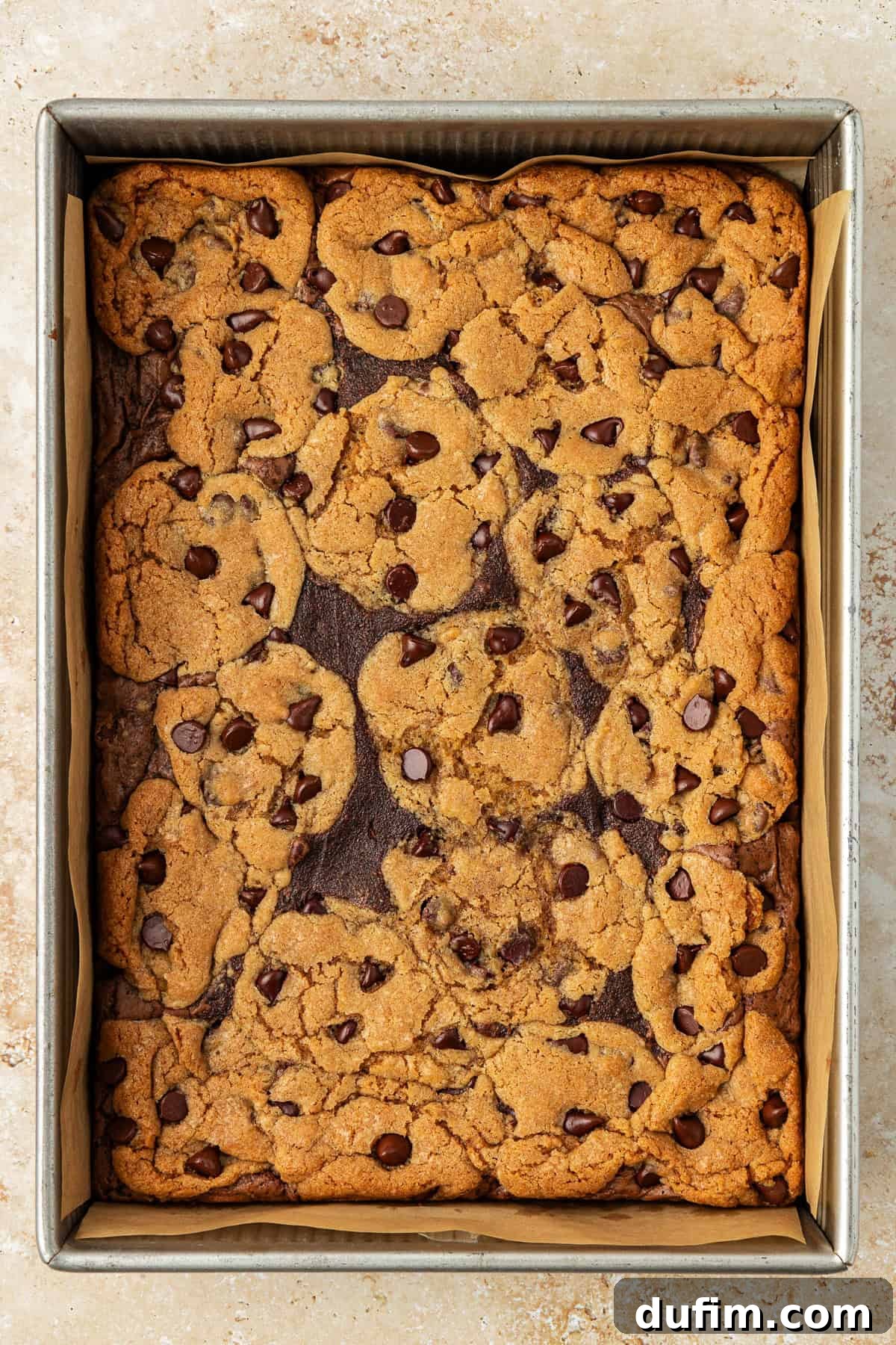 An overhead view of freshly baked, warm brookies in a metal baking pan lined with parchment paper, showcasing their golden chocolate chip cookie tops and rich brownie bases, ready to be cooled and sliced into perfect bars.