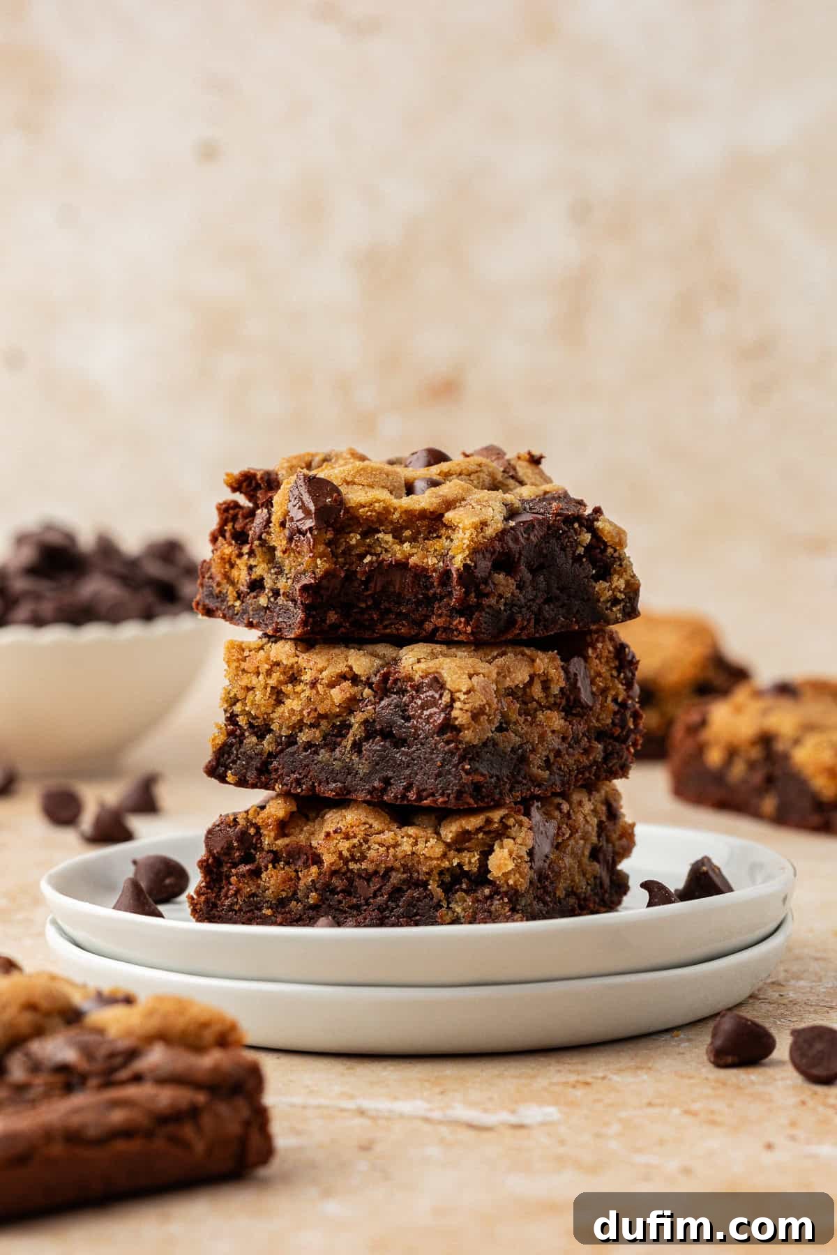 Three perfectly baked brookies stacked in a tower on top of two small white plates, with more brookies and loose chocolate chips scattered artfully on a light surface, ready for serving or further customization.
