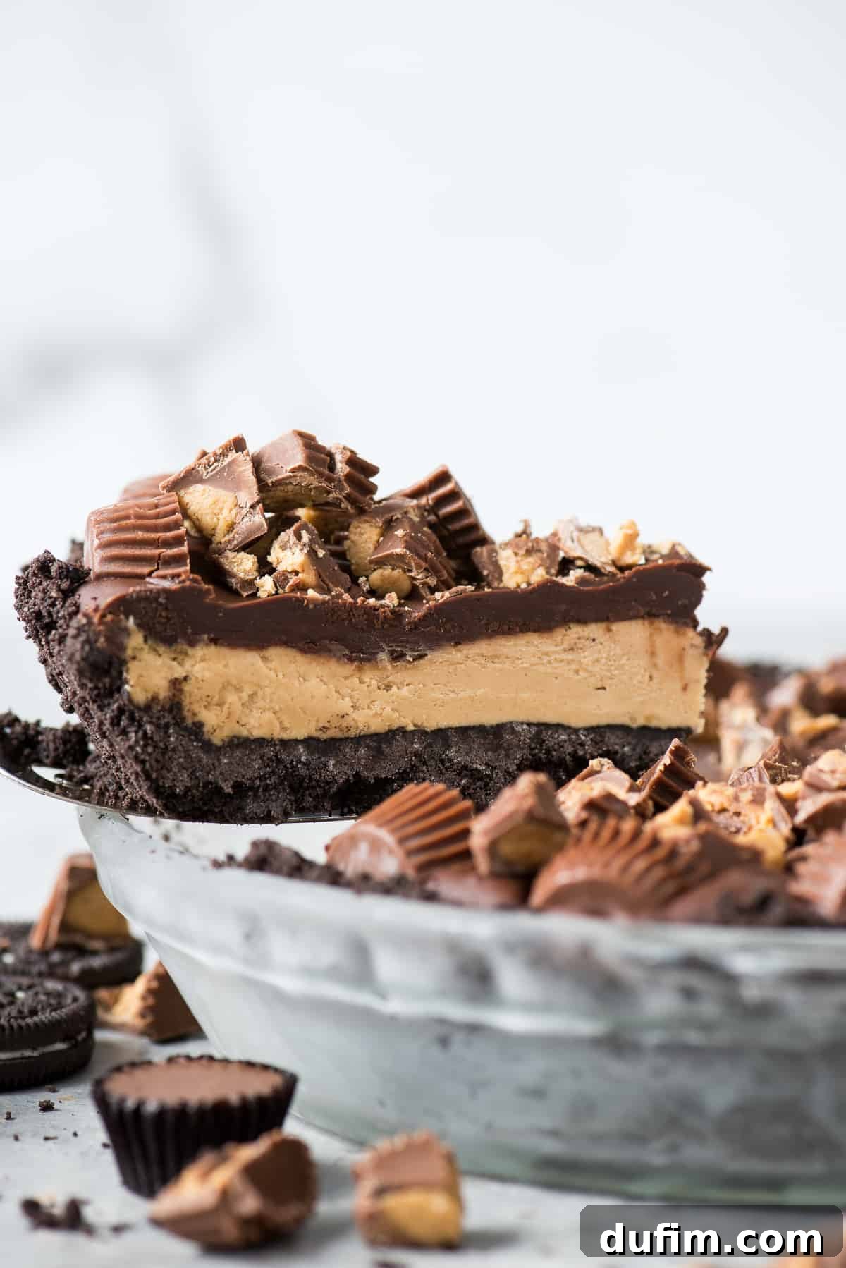 A delectable slice of Reese's Peanut Butter Cup Pie being lifted from a glass pie plate, showcasing its distinct layers of rich chocolate crust, creamy peanut butter filling, and luscious chocolate ganache, adorned with chopped Reese's candies.