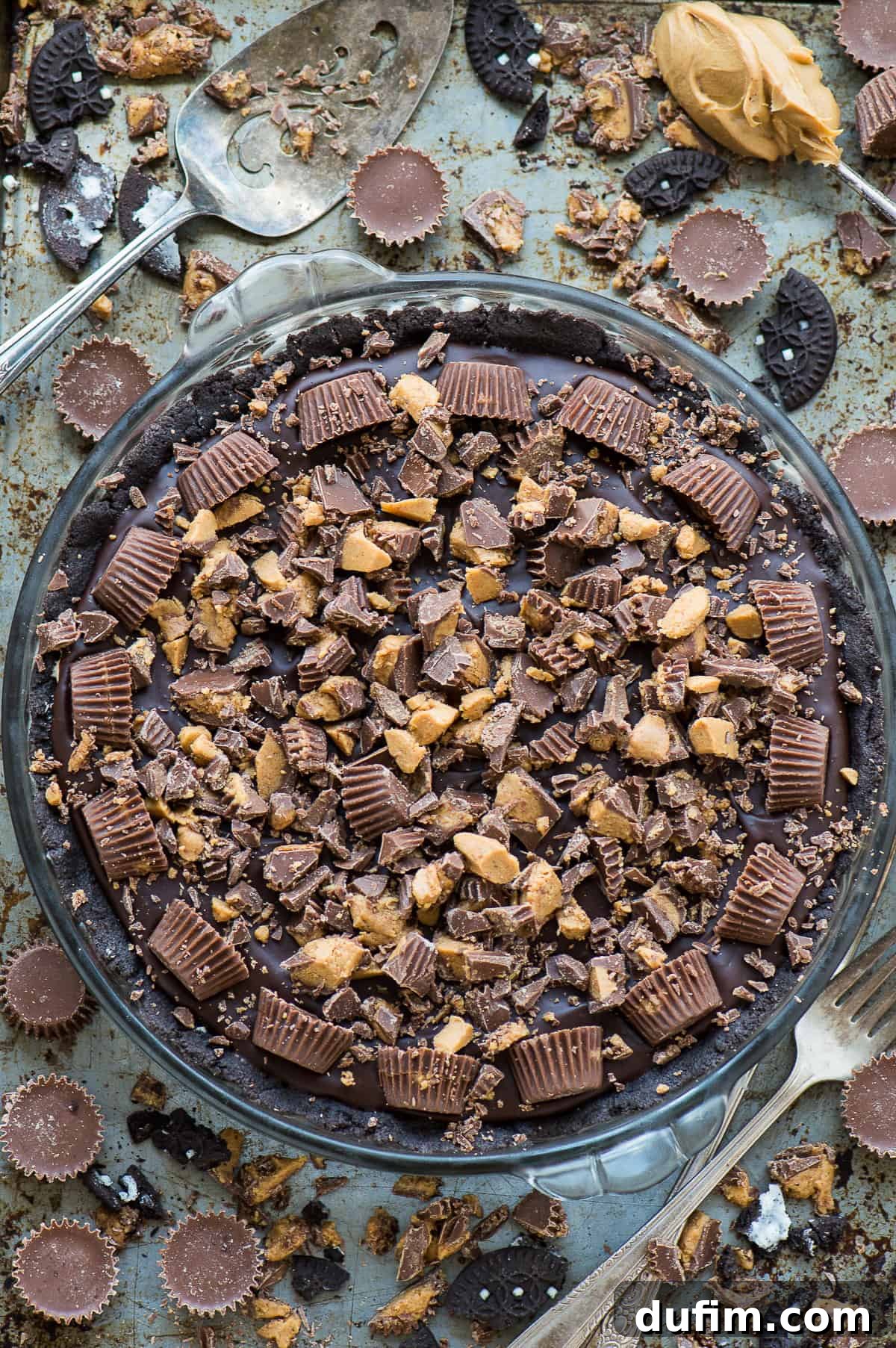 A whole chocolate peanut butter pie, generously topped with an assortment of Reese's Peanut Butter Cups, rests in a clear glass pie pan on a rustic metal background, ready to be sliced and served.